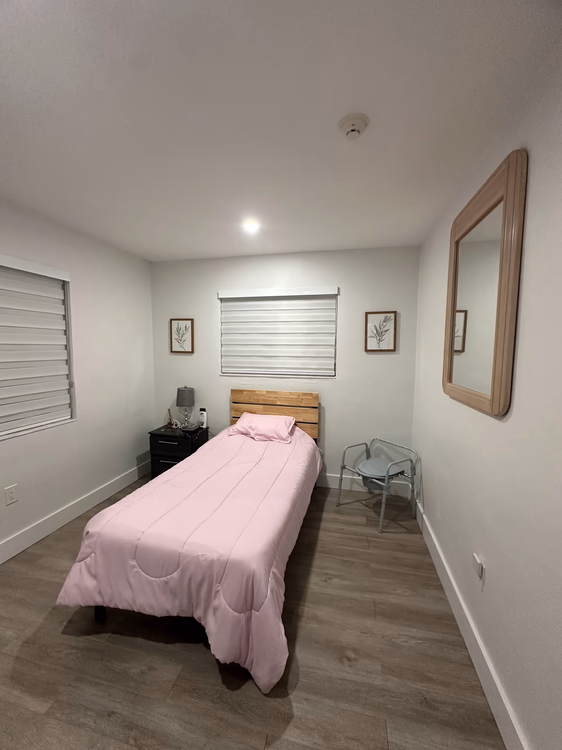Small bedroom with single bed, pink quilt, nightstand, window blinds, wall mirror, and chair.