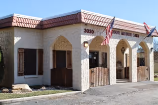 Exterior view of Windsor Mission Oaks facility showing a beige stucco building with a red tiled roof, arched entryways, two American flags, and the address number 3030 displayed near the entrance.