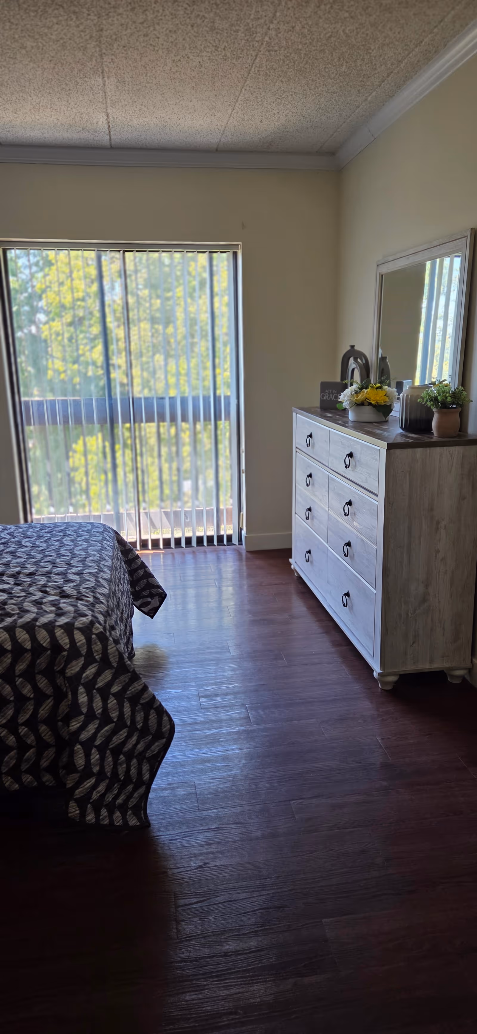 A bedroom with a bed, dresser, mirror, and sliding glass door with blinds.