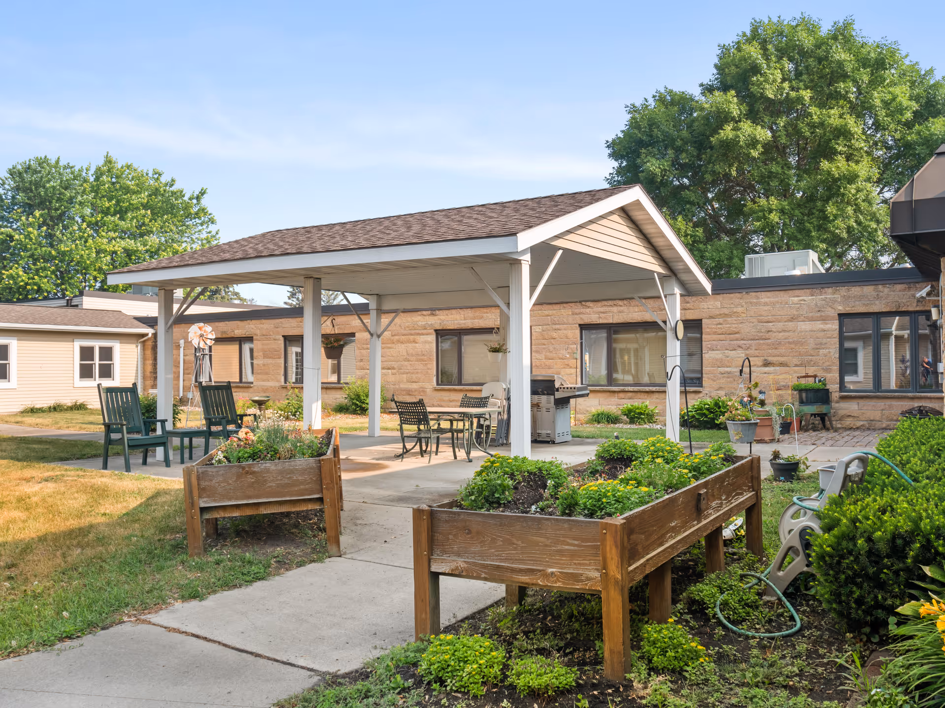 Outdoor patio area at Good Samaritan Society - Arlington - Fairview featuring a covered seating area with chairs and a table, surrounded by raised garden beds with plants and flowers. The building exterior is visible in the background with trees and a clear sky.