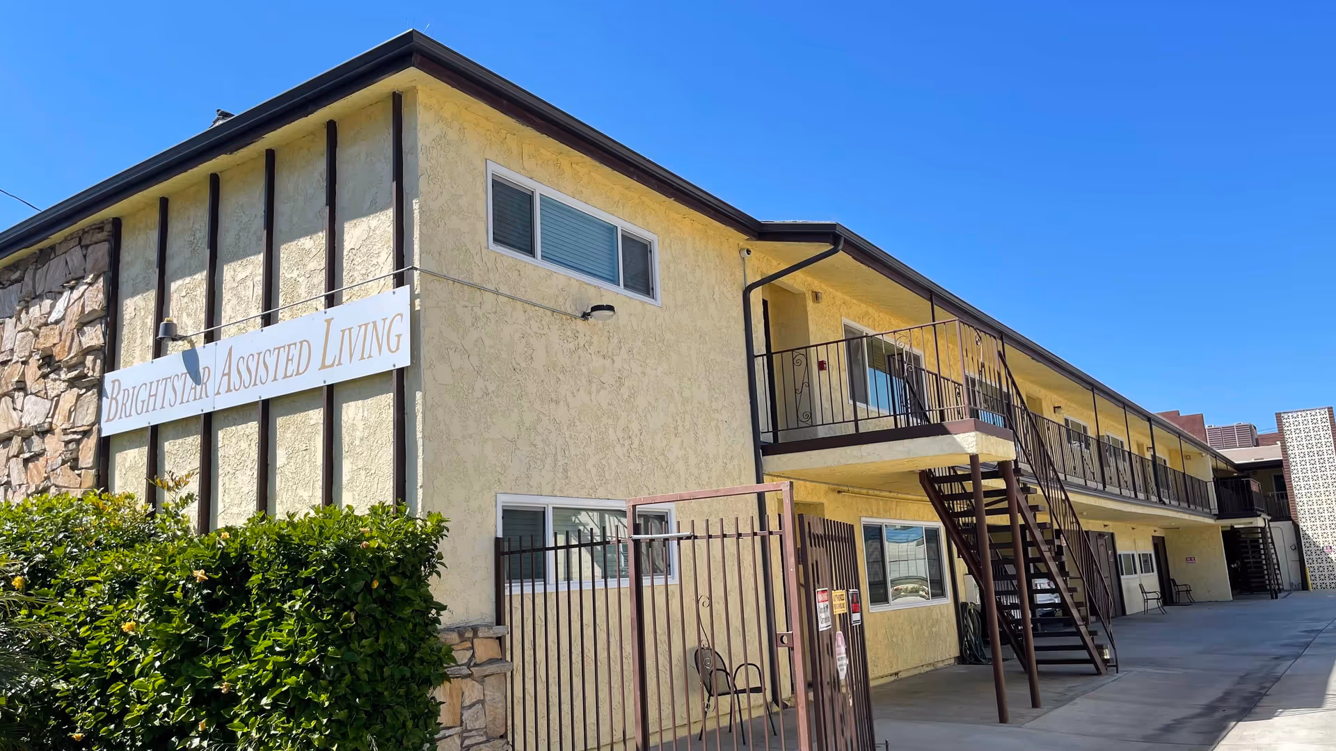 Exterior view of a two-story assisted living facility building with a yellow stucco facade and stone accents. The building has a sign that reads 'Brightstar Assisted Living'. There are metal railings and staircases leading to the upper floor, and a gated entrance with some greenery in front. The sky is clear and blue.