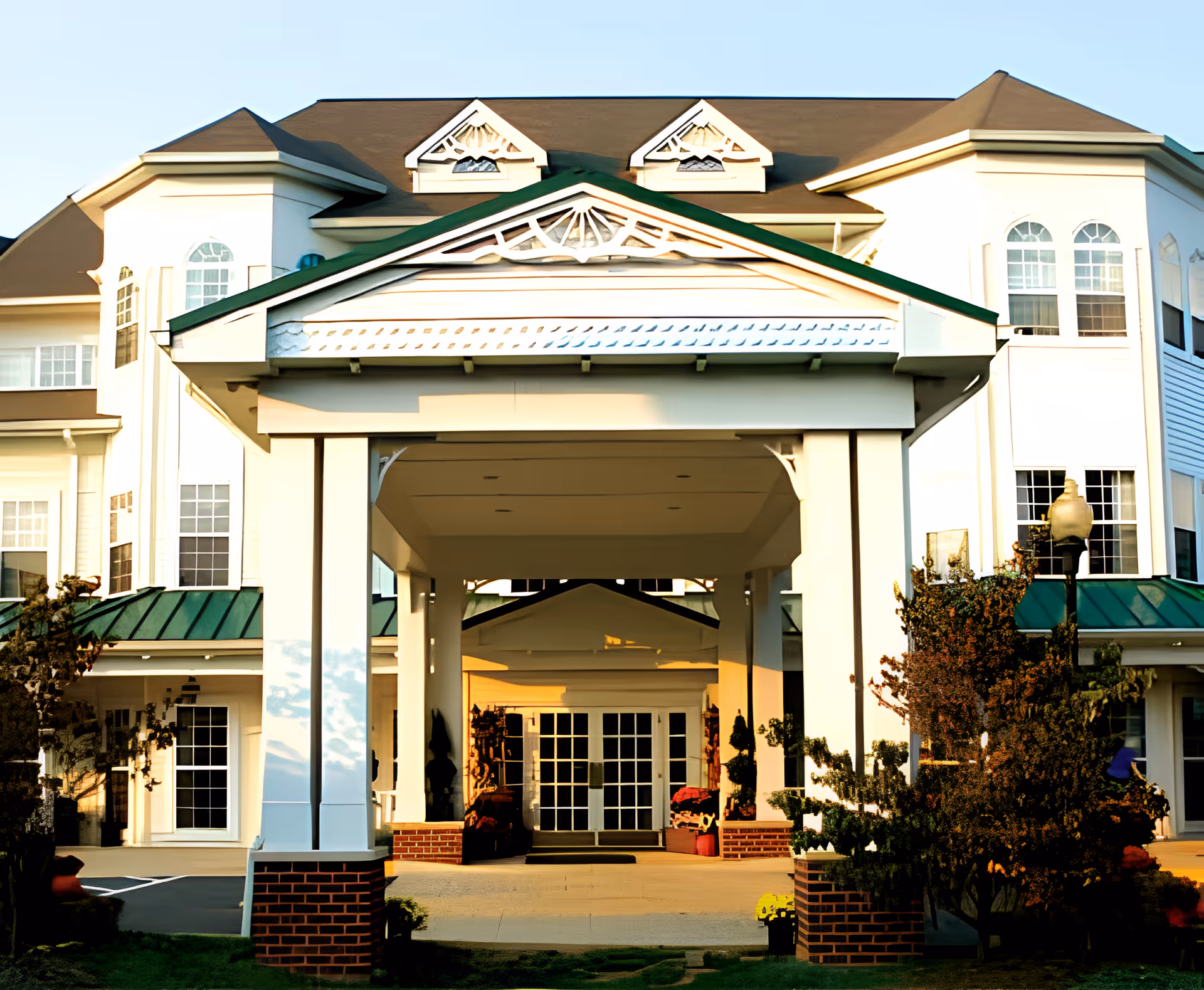 Front exterior view of a senior living facility named Spring Hills Somerset, featuring a large covered entrance with white columns and a green roof, surrounded by windows and landscaping.