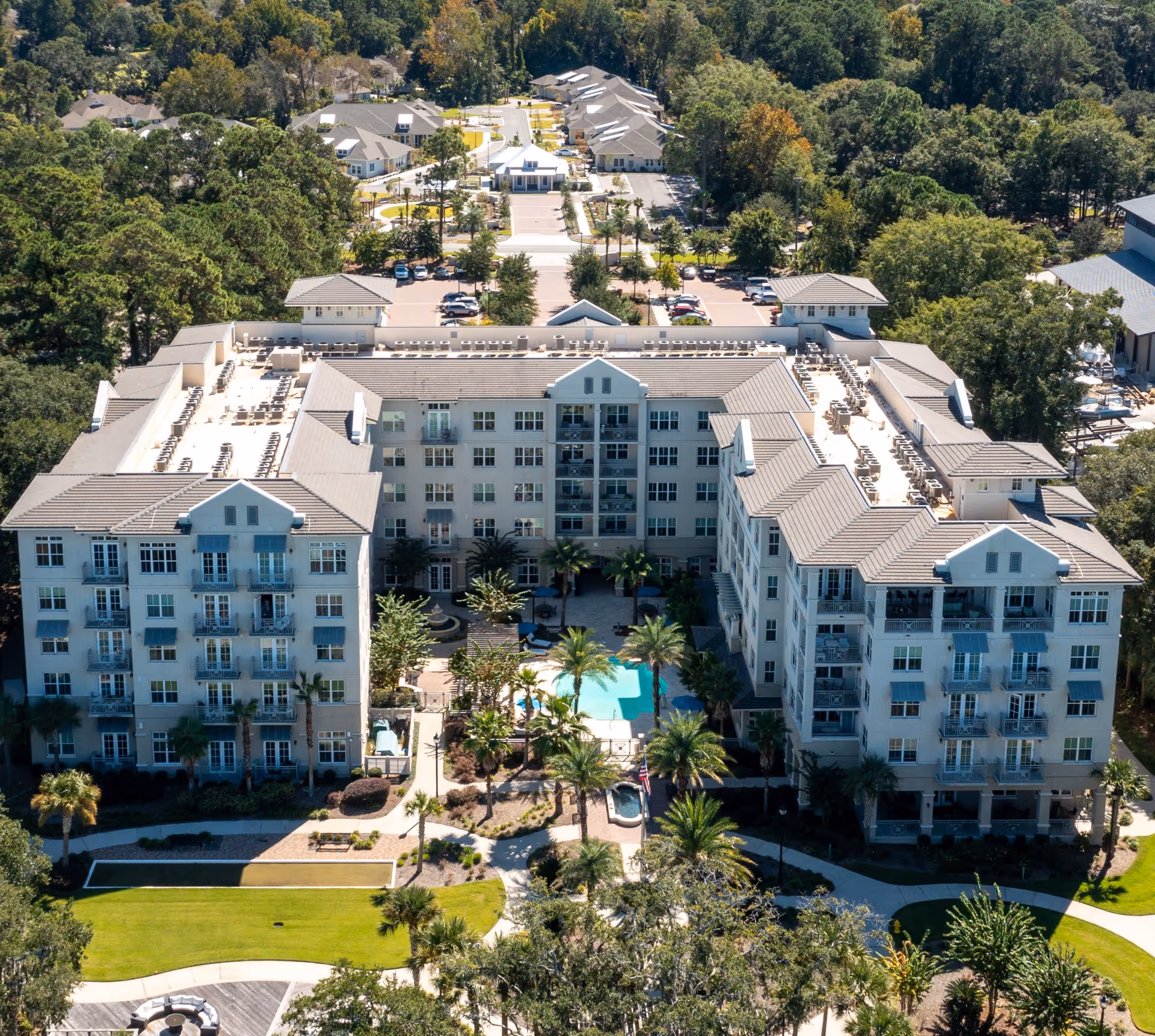 Aerial view of Bayshore on Hilton Head Island senior living facility showing a large multi-story building with balconies, a central courtyard with a swimming pool, palm trees, and landscaped gardens. Surrounding the building are parking areas, additional smaller buildings, and dense green trees.