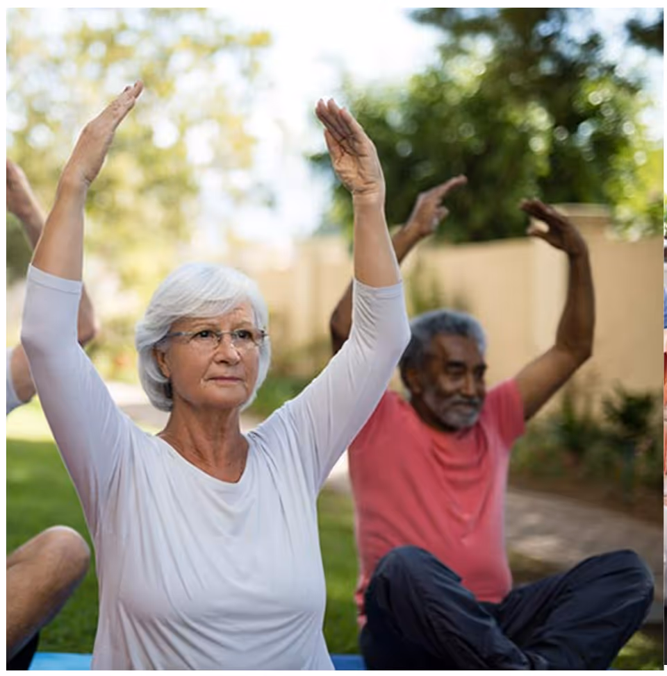 A group of elderly people sitting on yoga mats outdoors, raising their arms above their heads as part of a stretching or exercise routine. The setting is a garden or patio area with greenery and a beige wall in the background.