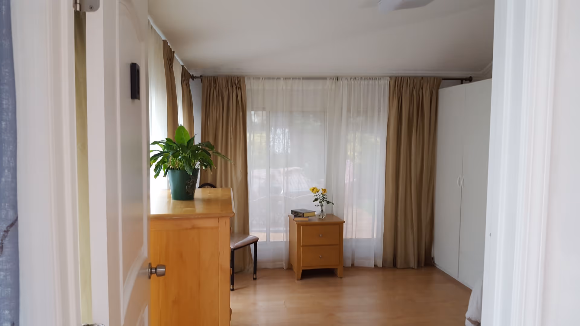 Small, tidy bedroom in a senior care facility with dresser, plant, and sheer curtains.