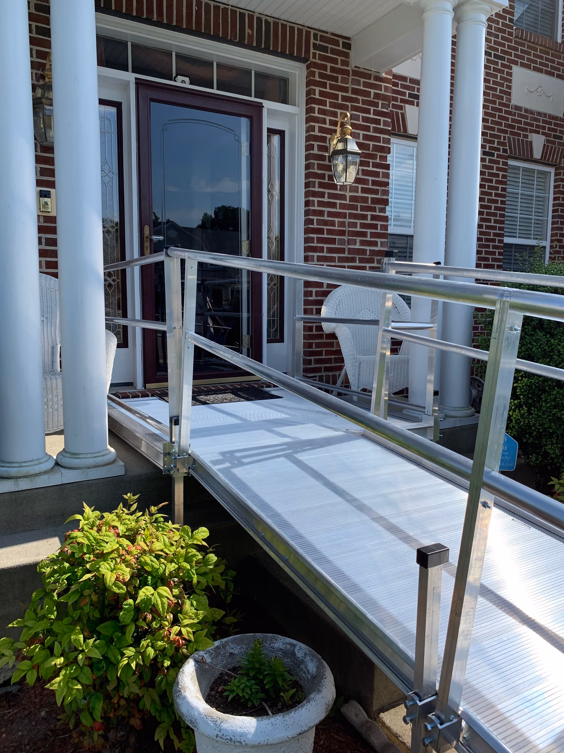 Exterior brick entrance with a metal ramp, porch seating, and greenery.