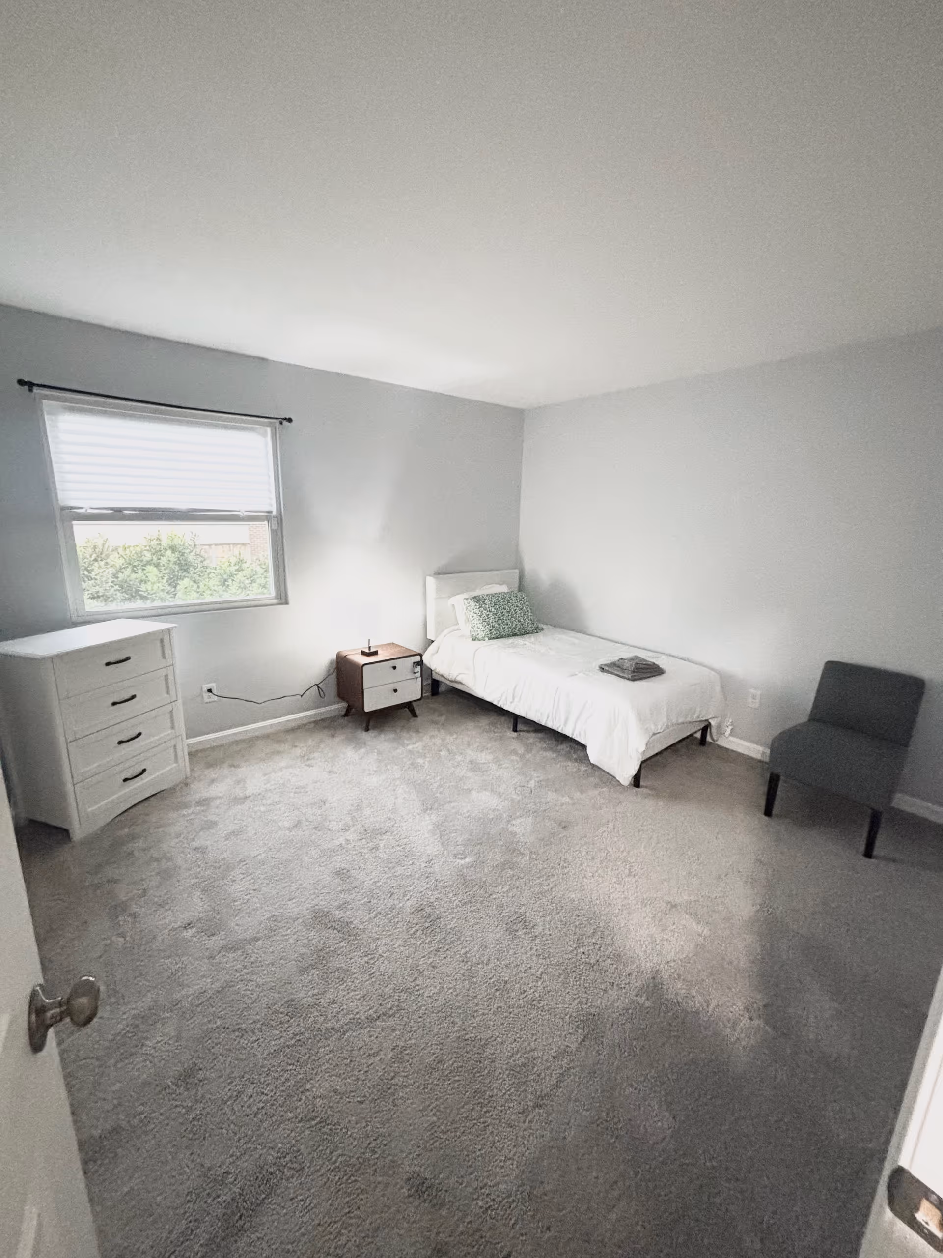 Bright, sparsely furnished bedroom with a bed, dresser, nightstand, and a gray chair.