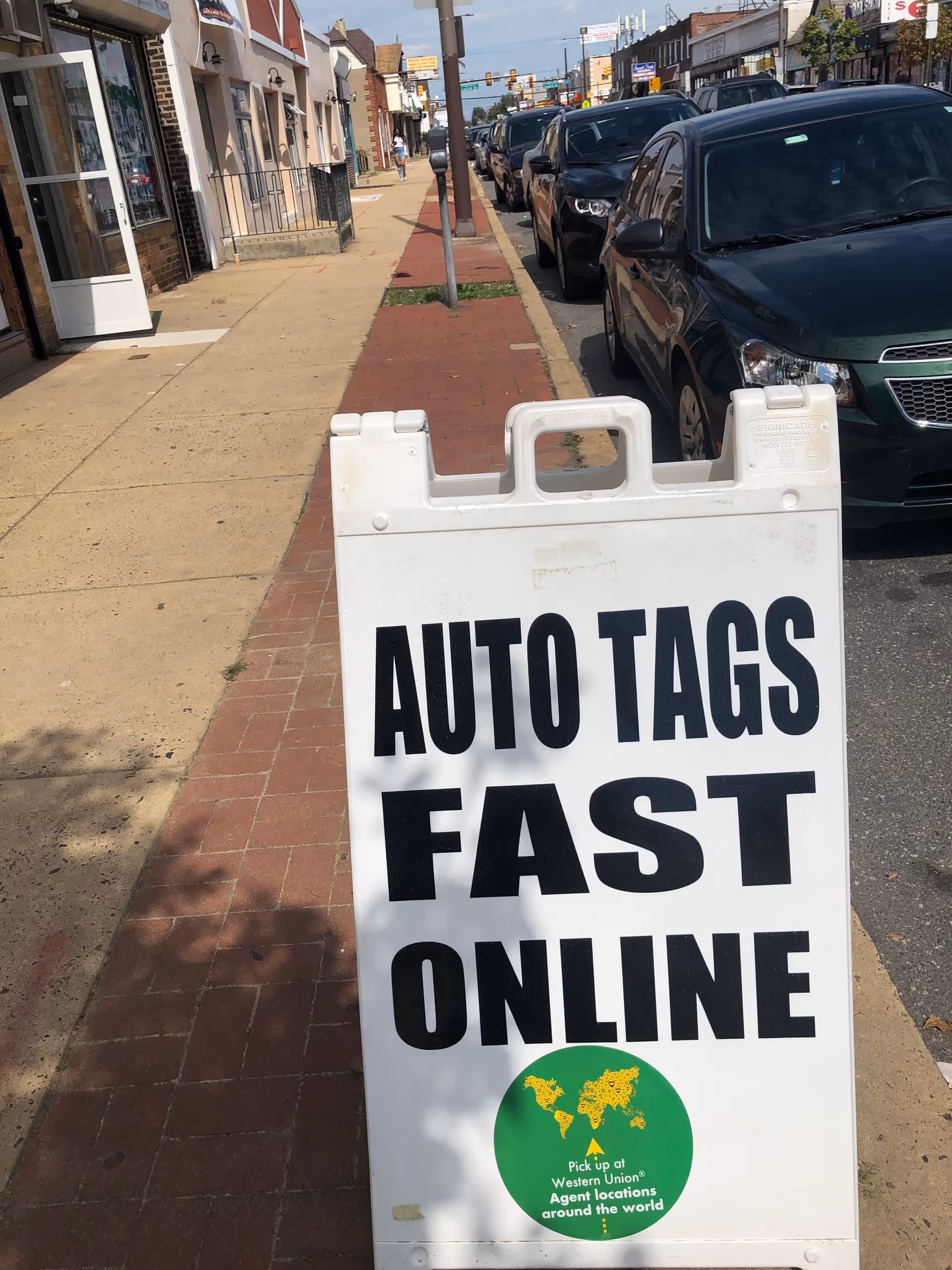 Sidewalk street with storefronts and a large white sign that reads AUTO TAGS FAST ONLINE.