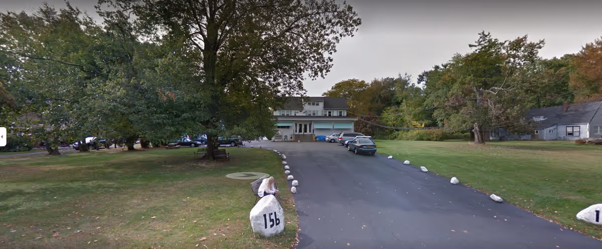Driveway lined with white-painted rocks leads to a two-story residential building set back on a grassy lot with large trees.