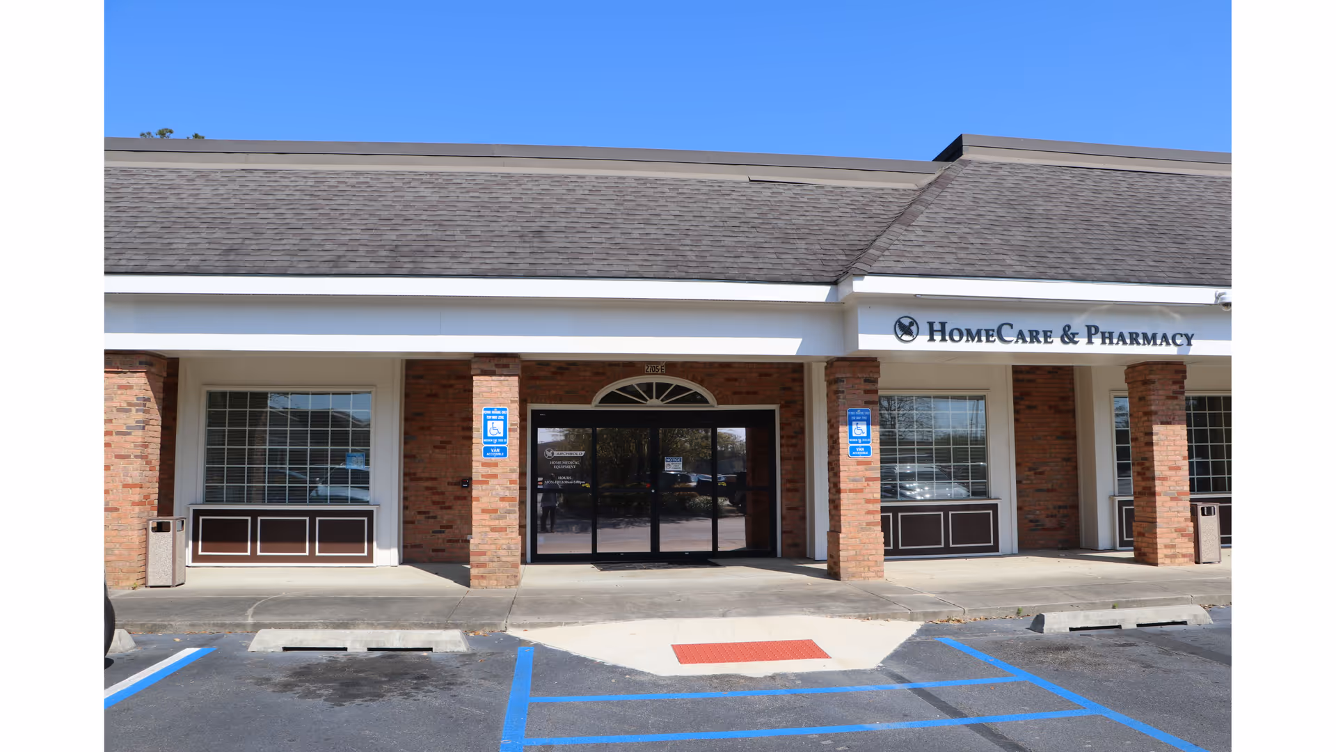Brick storefront with glass doors, pillars, and a HomeCare & Pharmacy sign under blue sky.