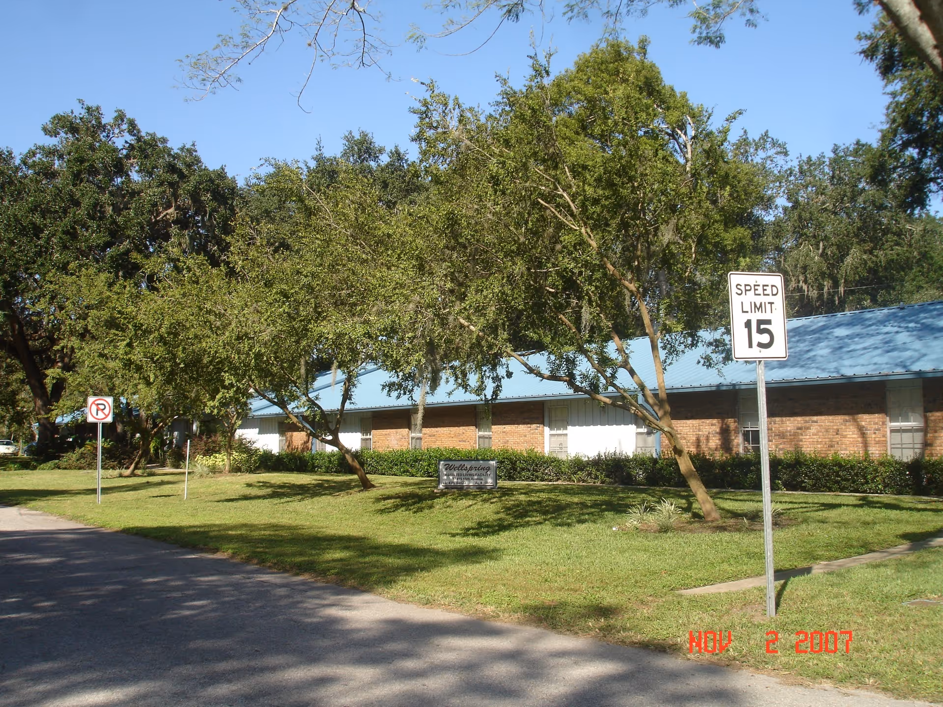 Exterior view of Wellspring Assisted Living facility showing a single-story brick building with a blue roof, surrounded by green grass and trees. There are traffic signs indicating a speed limit of 15 and no parking along the road in front of the building.