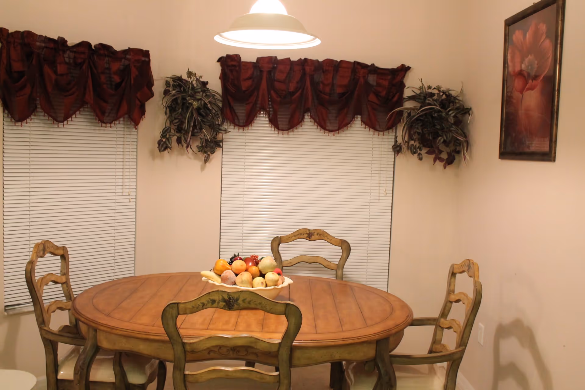 A dining room with a wooden oval table and four matching chairs. The table has a decorative bowl filled with artificial fruit. Two windows with closed blinds and dark red valances are on the wall behind the table, flanked by two hanging green plants. A framed floral painting is on the adjacent wall, and a ceiling light fixture hangs above the table.