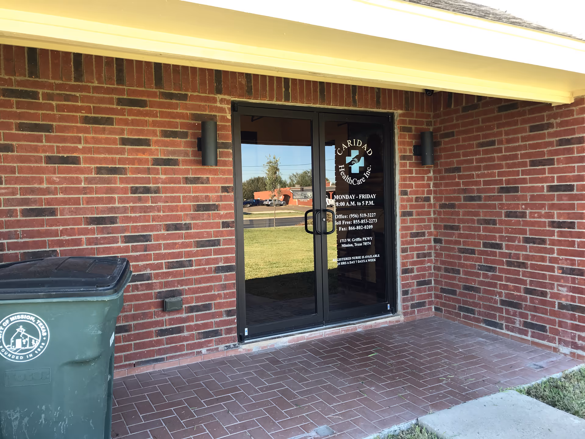 Brick exterior with a glass entrance door labeled Caridad Health Care Inc and a trash bin.