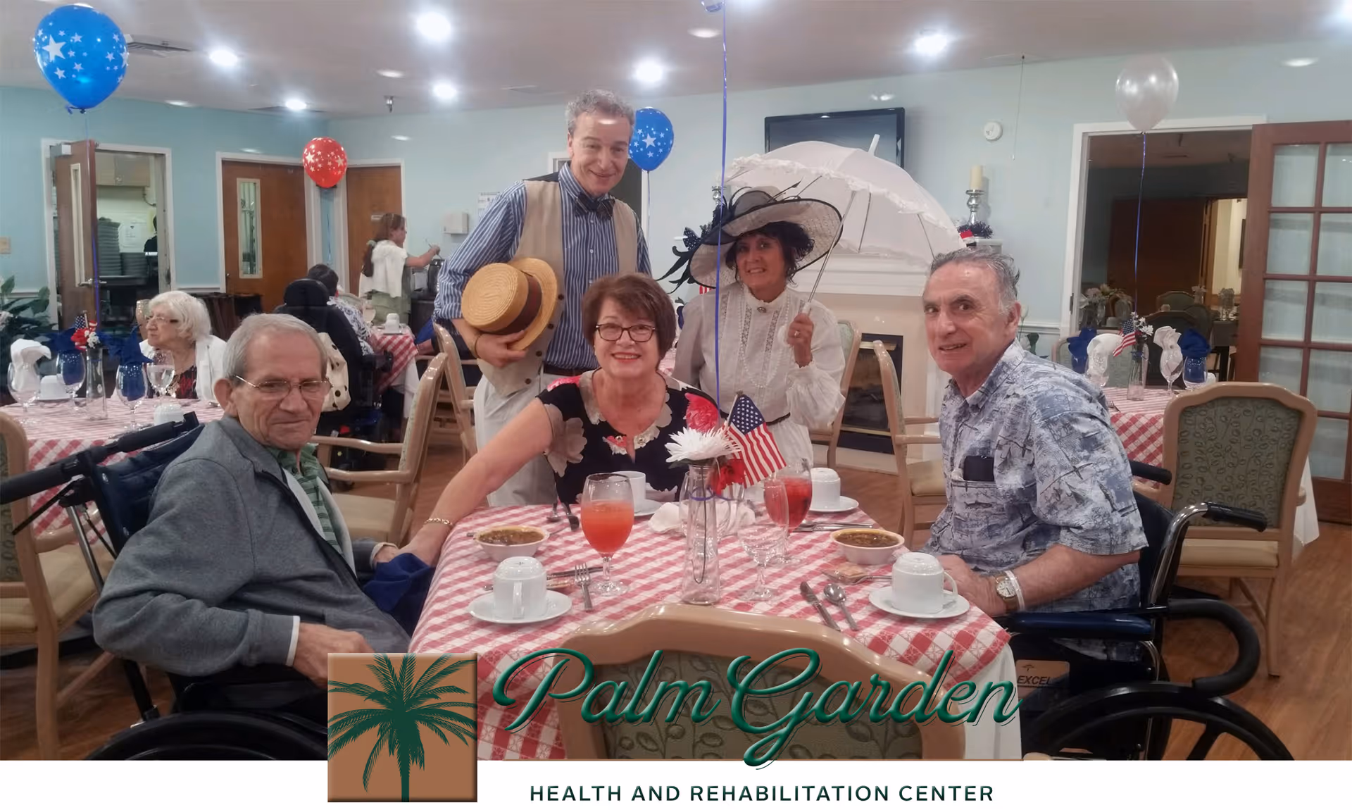 A group of elderly people and two staff members gathered around a table with a red and white checkered tablecloth in a dining room decorated with red, white, and blue balloons. The table is set with cups, glasses of red drink, and small American flags. One woman is holding a white parasol and wearing a vintage-style hat. The setting appears festive and cheerful.