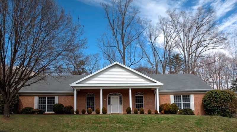 Front exterior of a single-story brick building with a white columned entrance, lawn, and leafless trees under a blue sky.