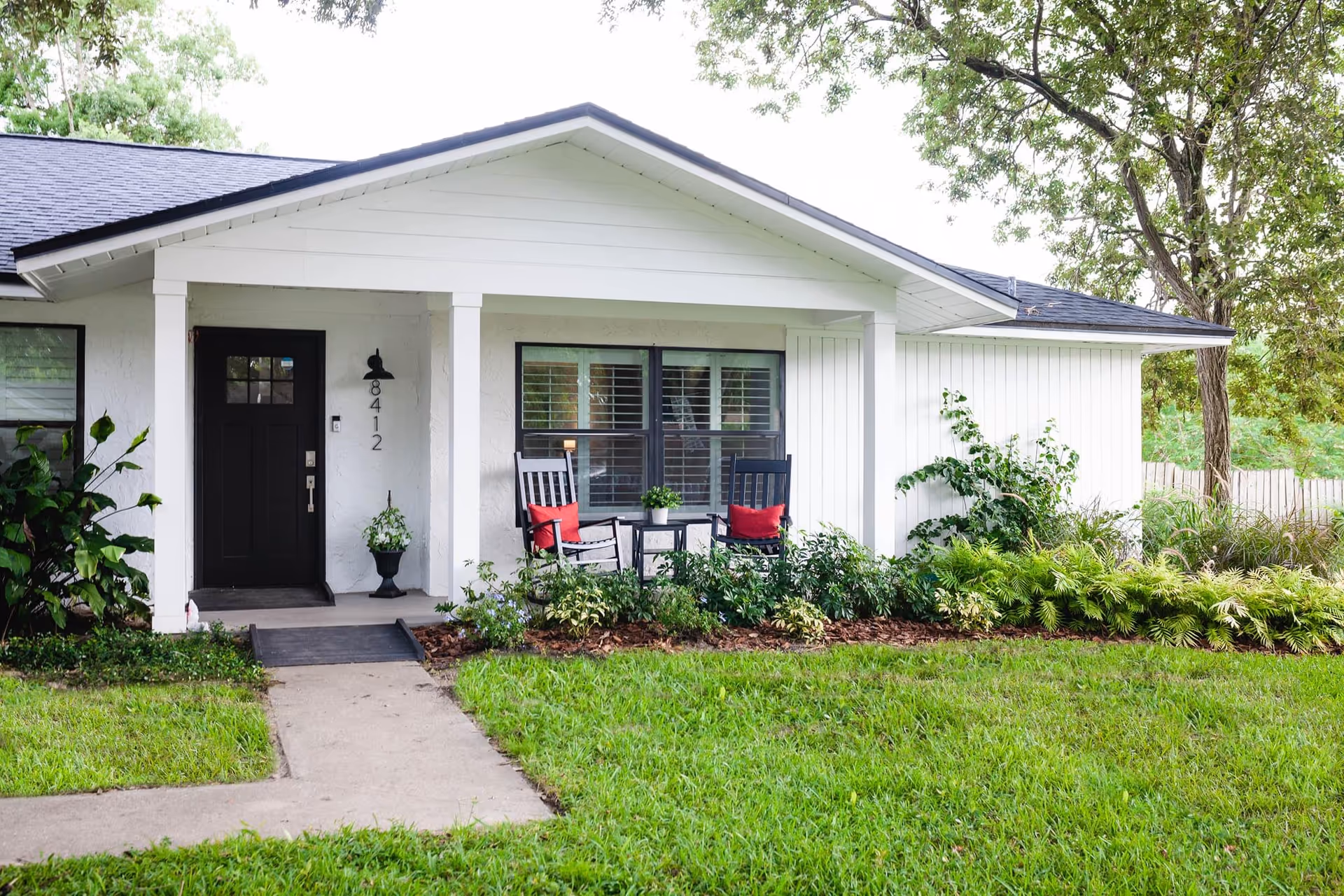 White single-story building with front porch, chairs, and lush landscaping.