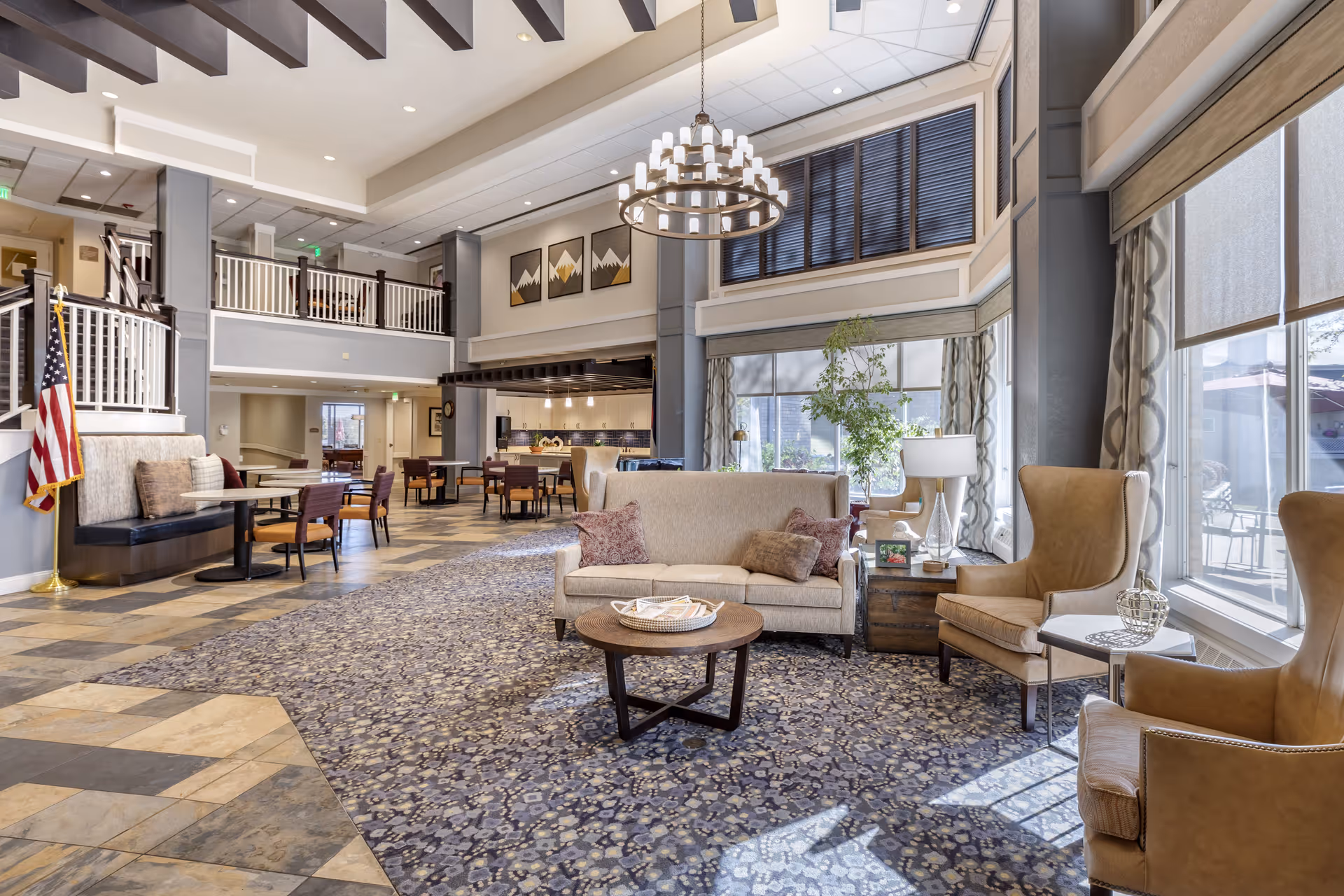 Spacious and well-lit senior living facility common area with a high ceiling, large windows, and a chandelier. The room features a beige sofa with pillows, two beige armchairs, a round wooden coffee table, and several dining tables with chairs. There is an American flag on a stand near a staircase with white railings, and decorative artwork on the walls. The floor is a combination of patterned carpet and tile.