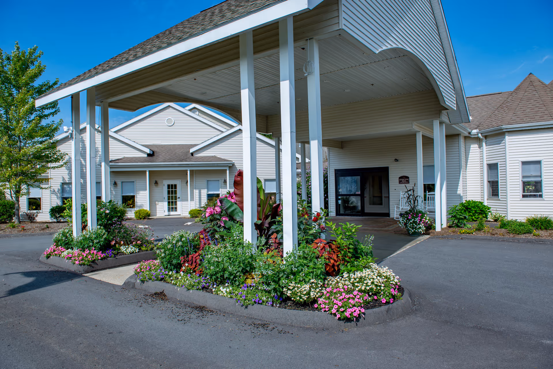 Entrance of Kimball Farms facility with a covered driveway supported by white pillars, surrounded by colorful flower beds and greenery under a clear blue sky.