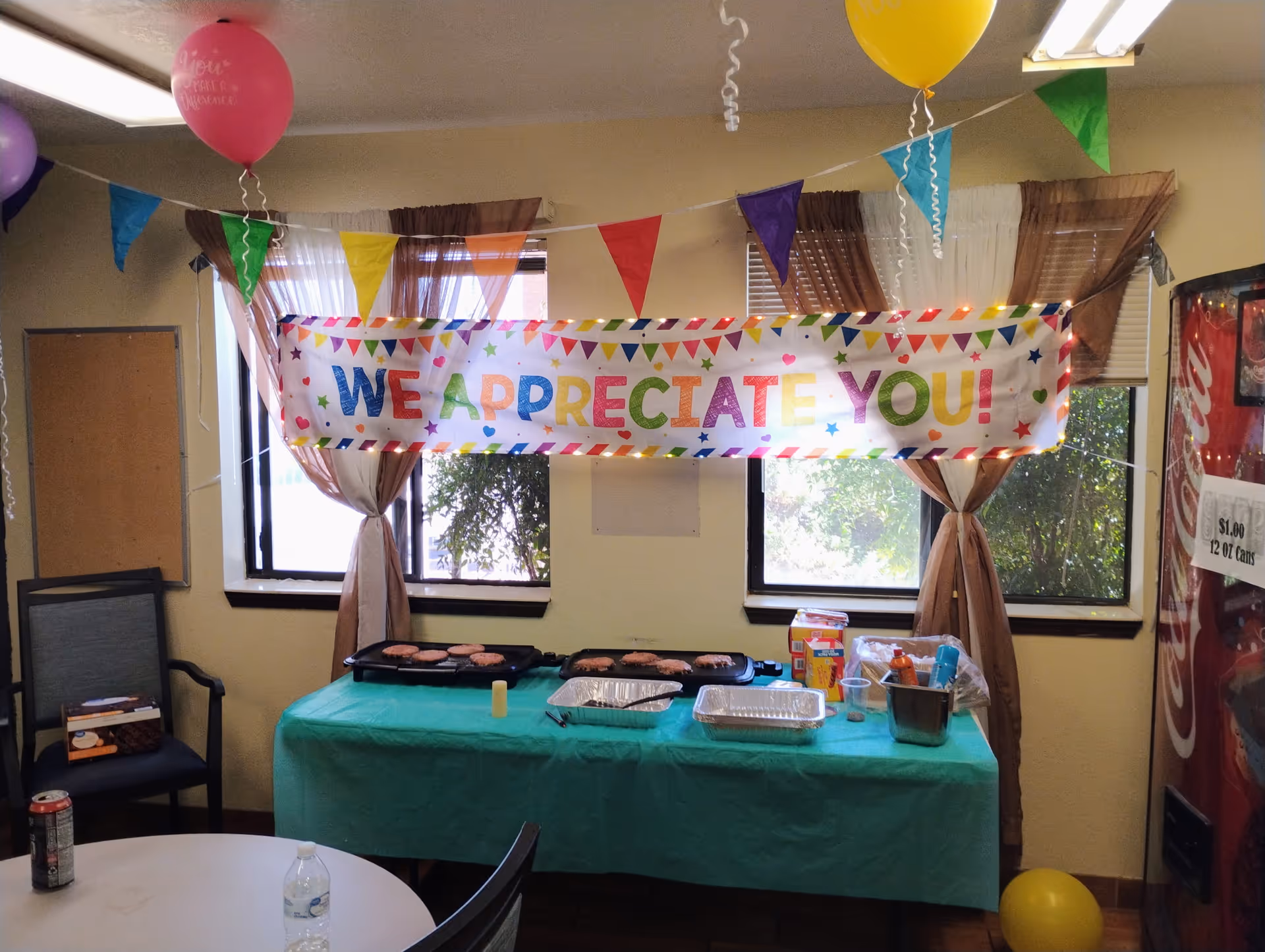 Indoor celebration setup with a long table, banner, balloons, and food trays.