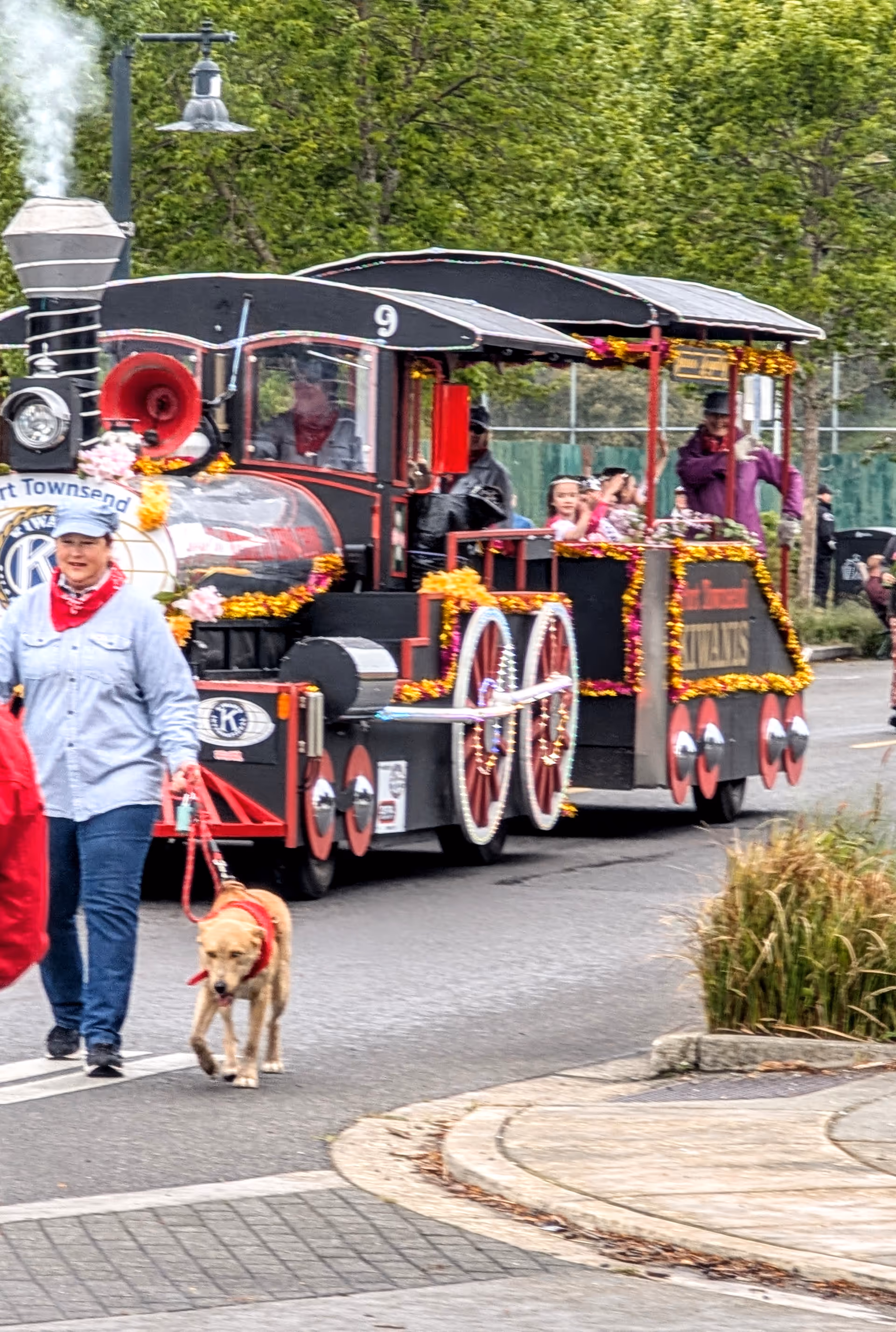 Outdoor street parade features a decorated miniature locomotive with passengers, while a person walks a dog.