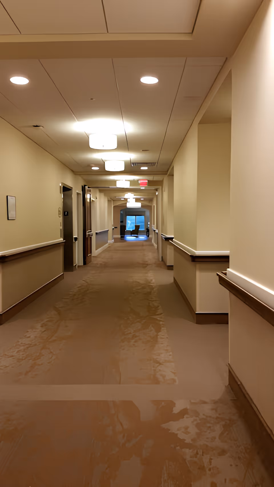 A long, empty hallway in a rehabilitation and nursing center with beige walls, brown carpet, handrails on both sides, and ceiling lights. At the end of the hallway, there is a window with a chair positioned in front of it.