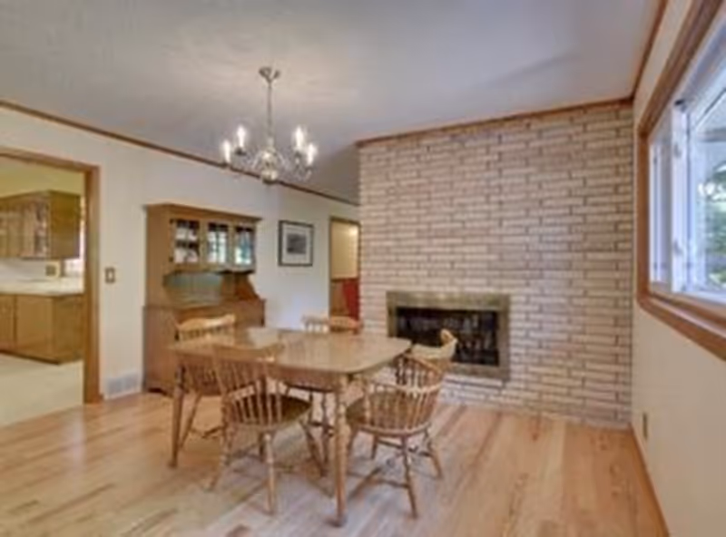 A wooden dining table with chairs in a room featuring a brick fireplace, hardwood floors, a chandelier, and a large window.