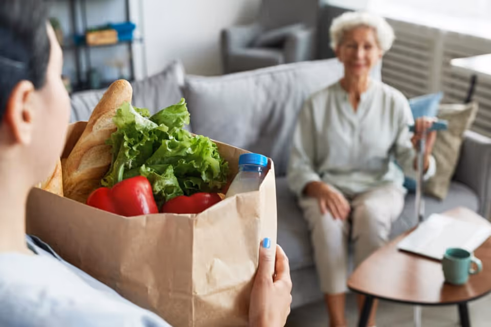 Person in foreground holds a grocery bag while resident sits on sofa indoors.