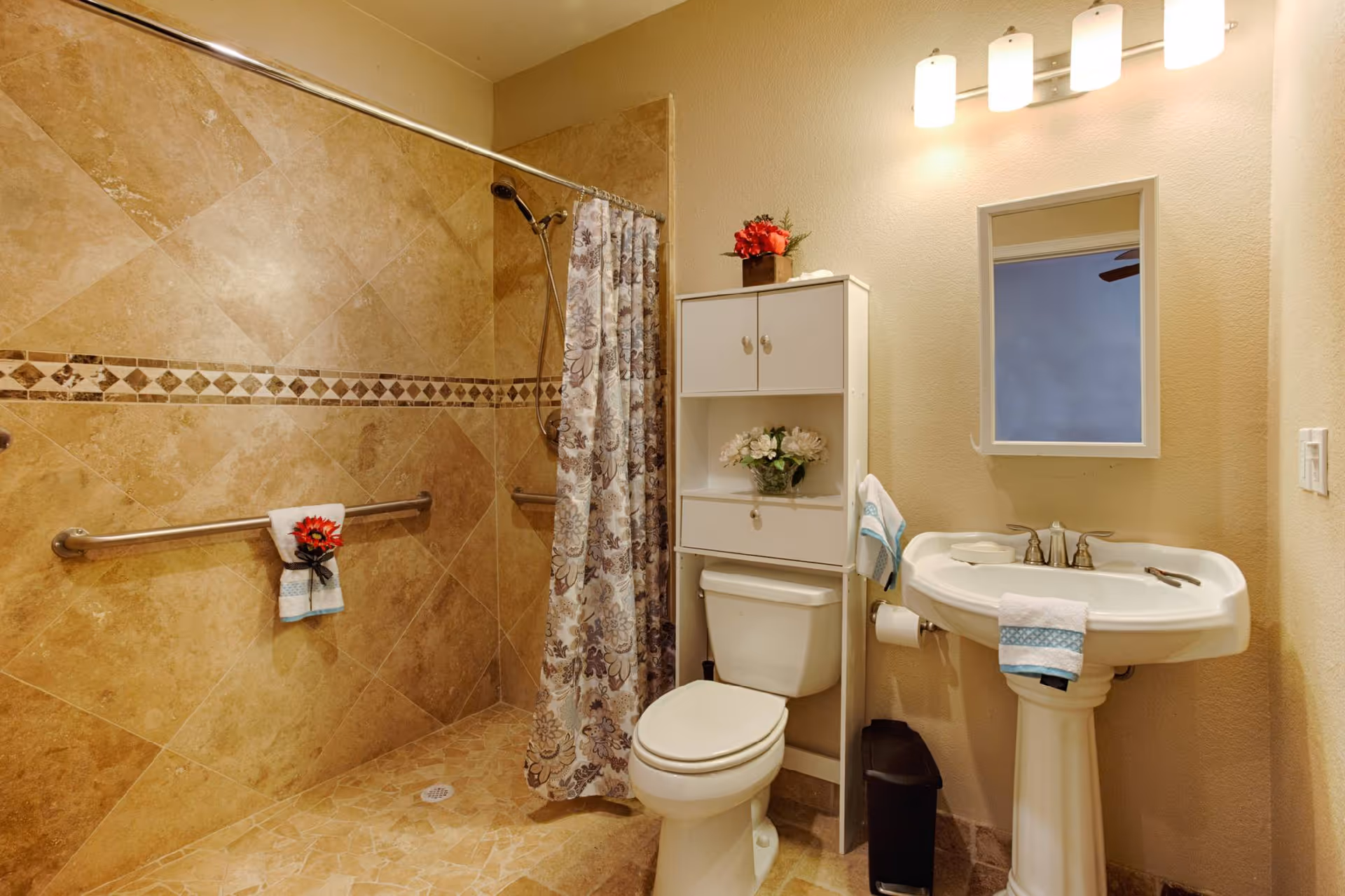 A bathroom with beige tiled walls and floor, featuring a walk-in shower with a floral shower curtain and grab bars. There is a white toilet with a white cabinet above it holding decorative flowers. A pedestal sink with a mirror above it and a light fixture with four lights is on the right side. Towels with blue accents and a red flower decoration are placed on the grab bar, sink, and cabinet.