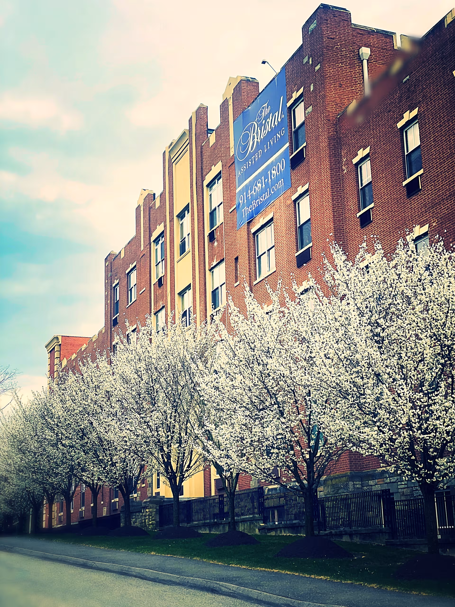 Brick building with The Bristal Assisted Living banner and blooming trees along sidewalk.