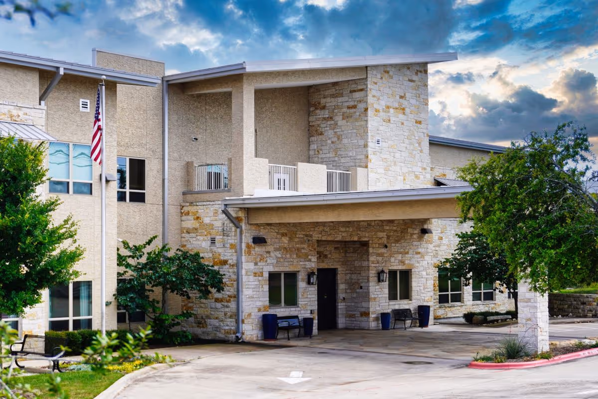 Stone and stucco senior living building entrance with a covered drive, American flag, benches, and trees under a cloudy sky.