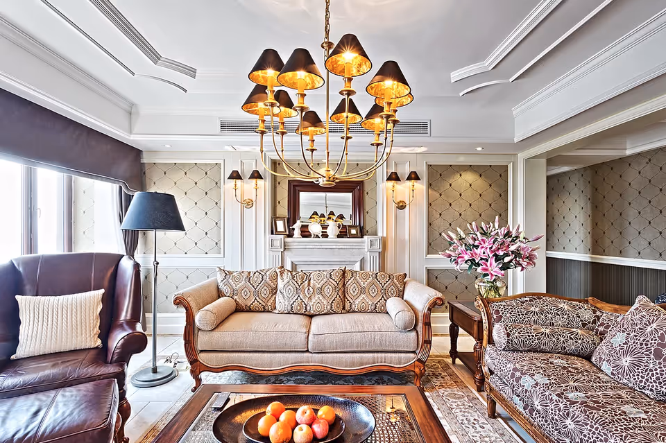A well-decorated living room with a beige sofa adorned with patterned cushions, a brown leather armchair with a white pillow, a floral-patterned couch, a wooden coffee table with a bowl of fruit, a floor lamp, a chandelier with multiple lampshades, and a fireplace with decorative items and a mirror above it.