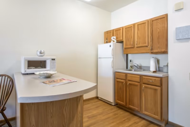Residential kitchenette with wooden cabinets, refrigerator, microwave, sink, and curved counter.