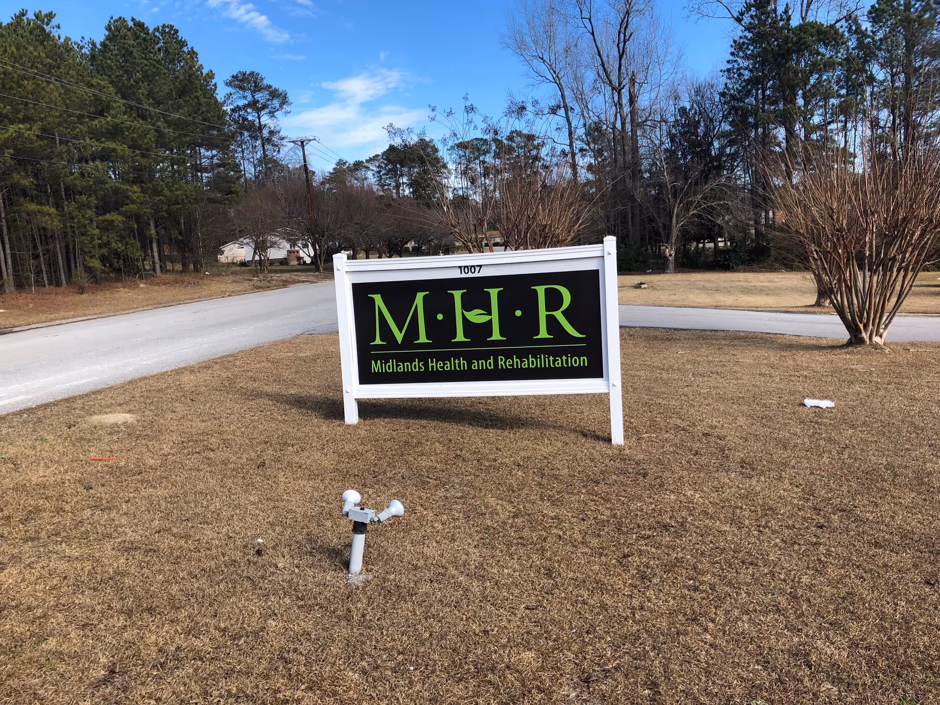 Outdoor view of a sign for Midlands Health and Rehabilitation Center placed on a grassy area near a road, with trees and a clear blue sky in the background.