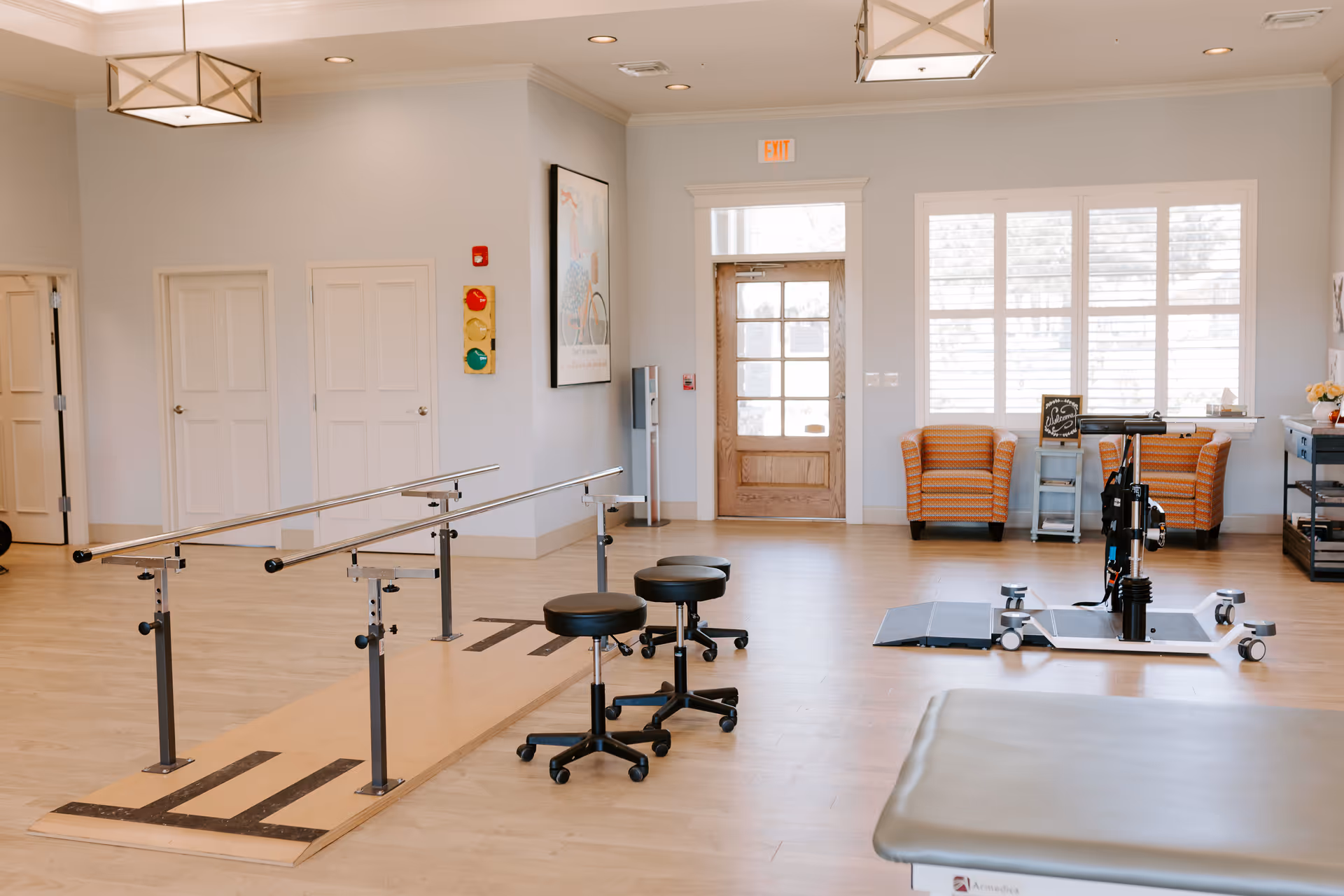 Rehabilitation gym area with parallel bars, chairs, and exercise equipment in a bright room.