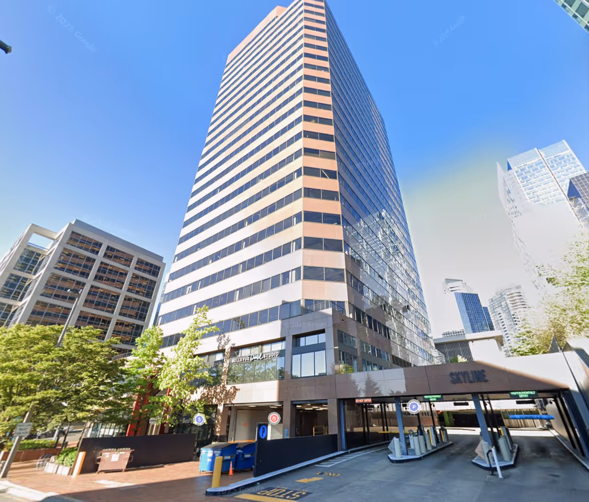 Tall glass skyscraper dominates street view with an entrance and nearby trees.
