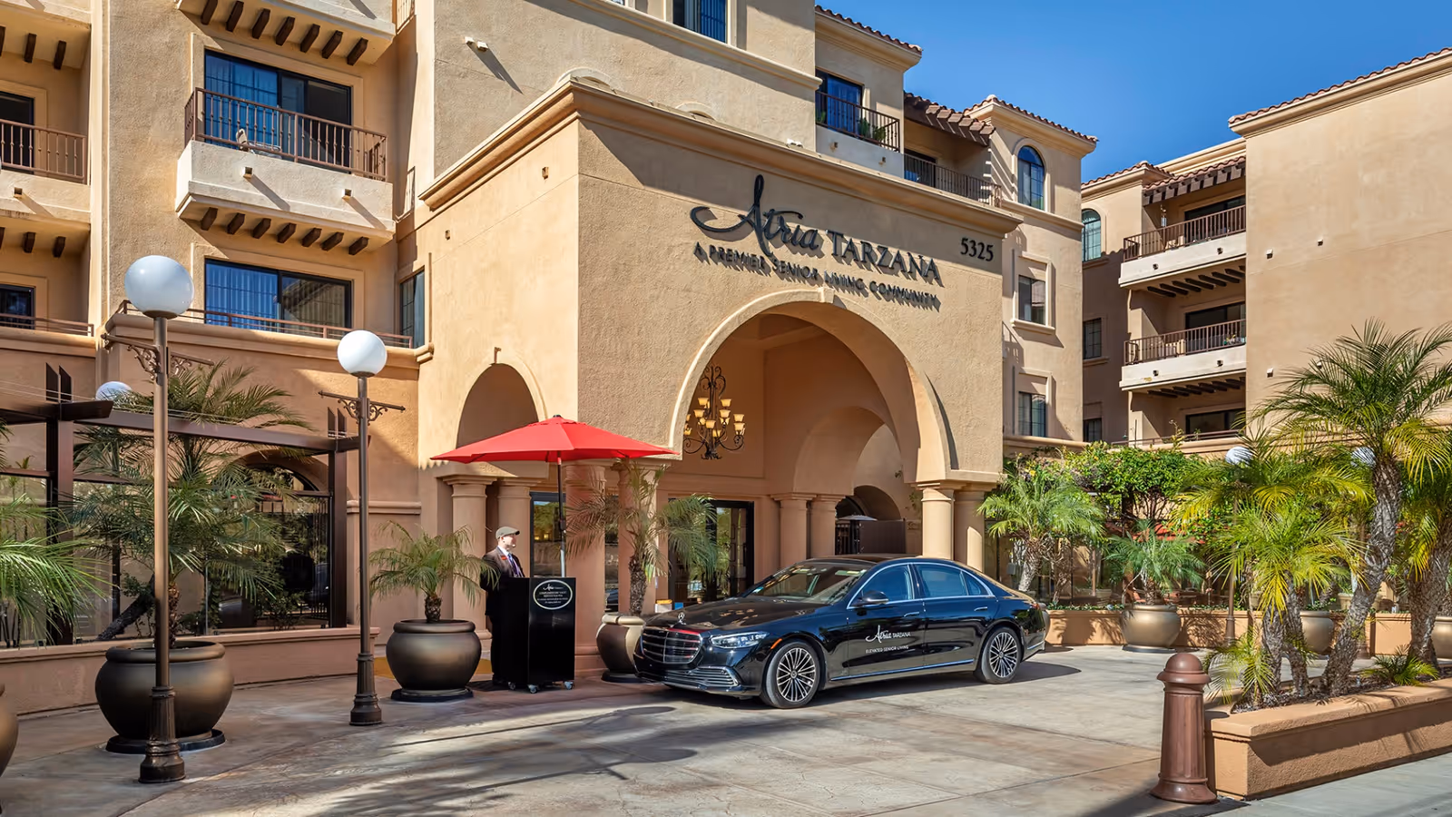 Atria Tarzana entrance with arches, beige stucco, palms, a red umbrella, and a parked black sedan.
