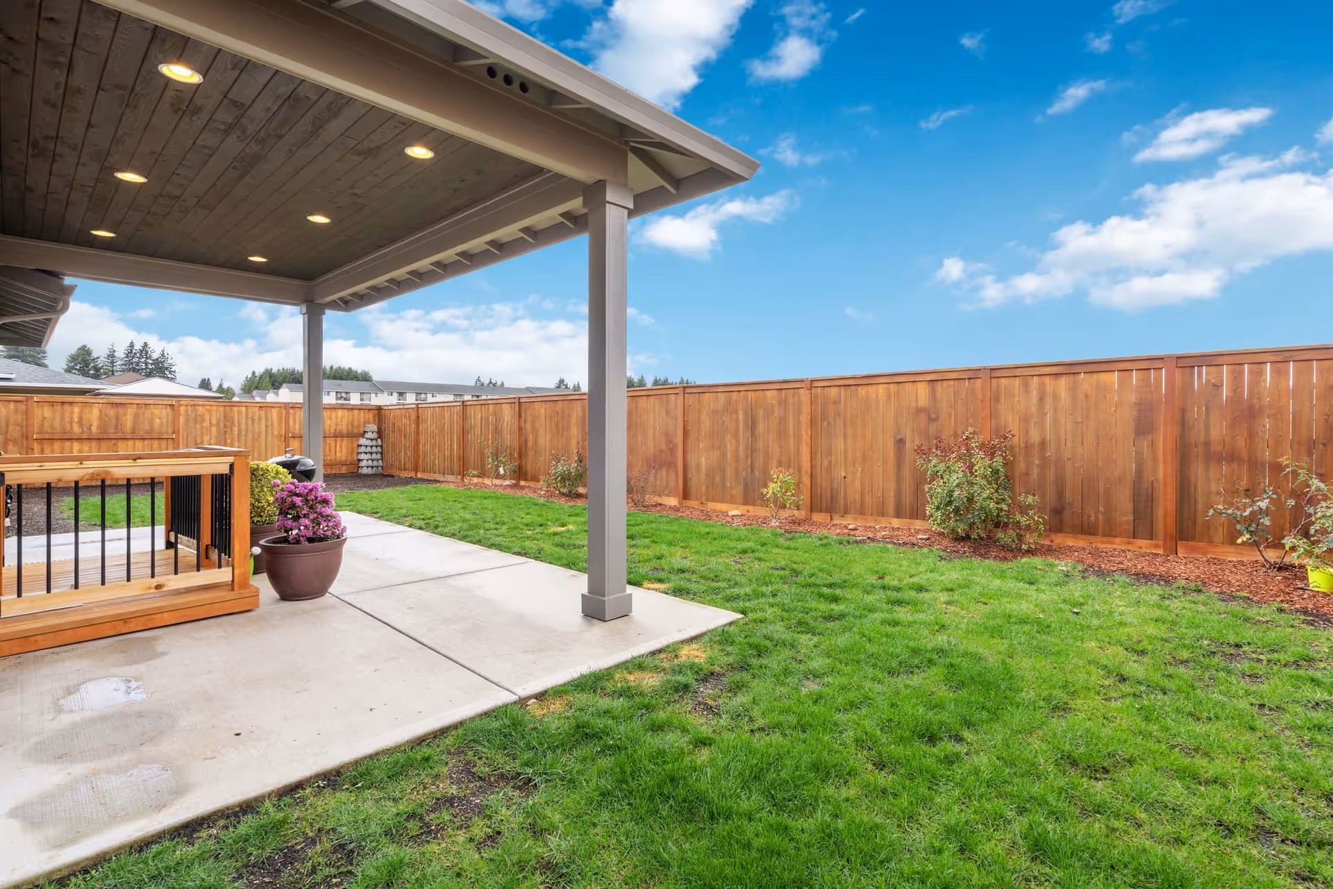 Covered patio with recessed lights overlooking a grassy backyard bordered by a tall wooden fence and potted plants.