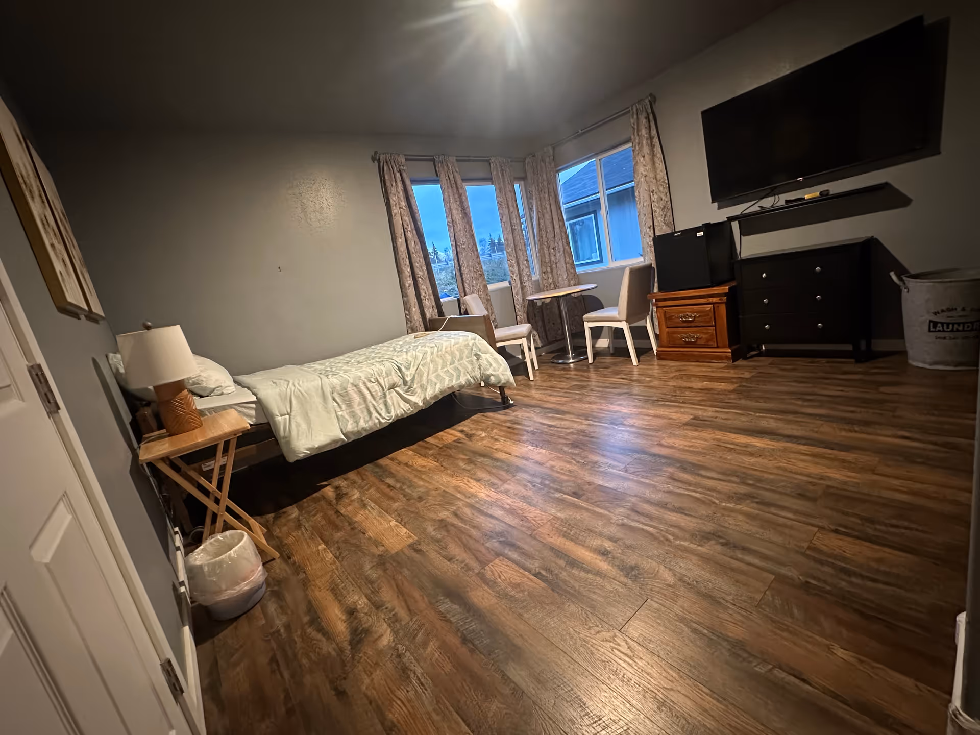 A furnished senior living bedroom with bed, dresser, TV, chairs, and windows.