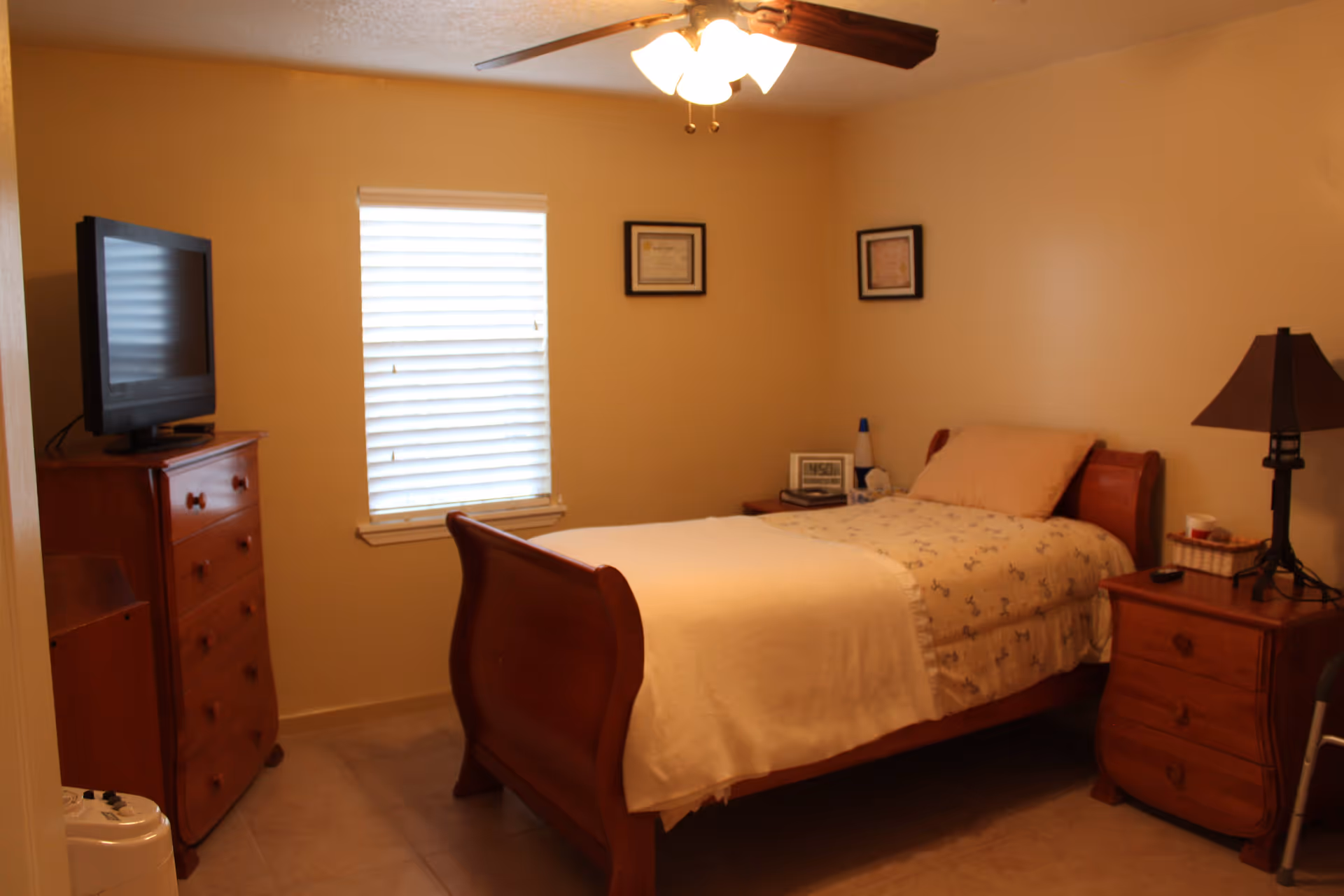 A cozy bedroom with a wooden bed frame, a single bed covered with a light-colored blanket and patterned sheets. There is a wooden nightstand with a lamp and some items on it to the right of the bed. A wooden dresser with a flat-screen TV on top is positioned against the left wall. The room has a window with closed blinds and two framed pictures hanging on the wall above the bed. A ceiling fan with lights is mounted on the ceiling.