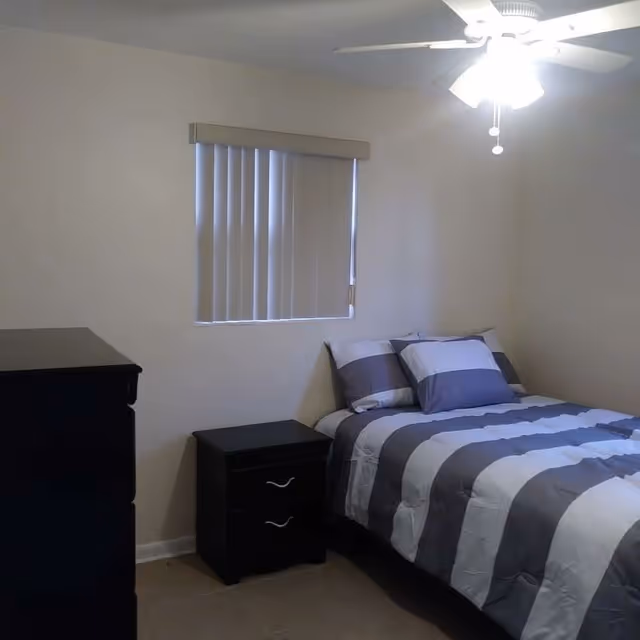 A small bedroom with a striped bed, nightstand, dresser, window blinds, and ceiling fan.
