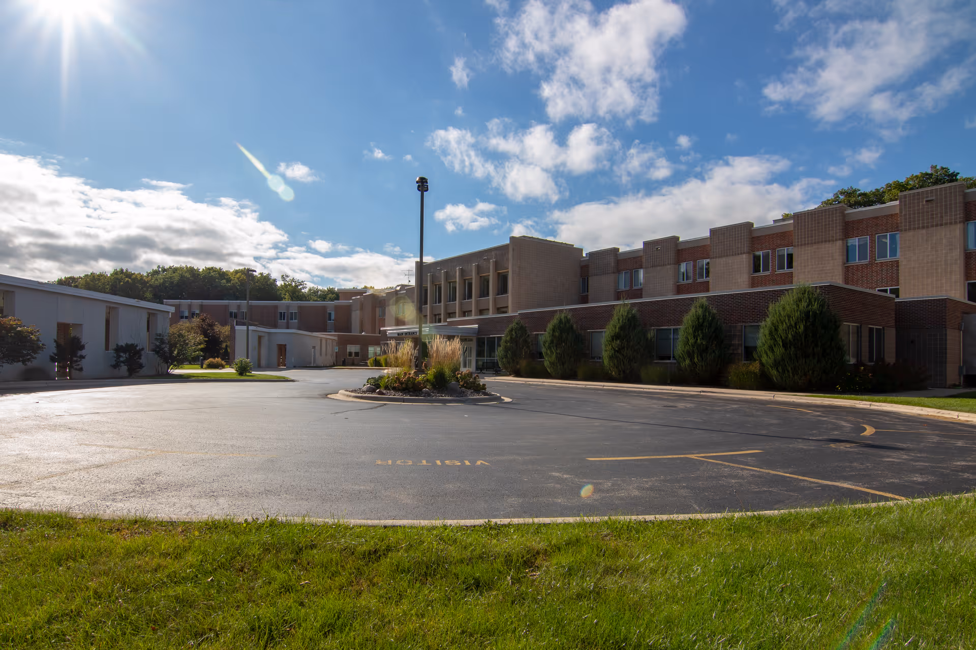 Exterior view of Rocky Knoll Health Care Center on a sunny day with a clear blue sky and some clouds. The building is a multi-story brick structure with several windows and a circular driveway in front, surrounded by green grass and some bushes.