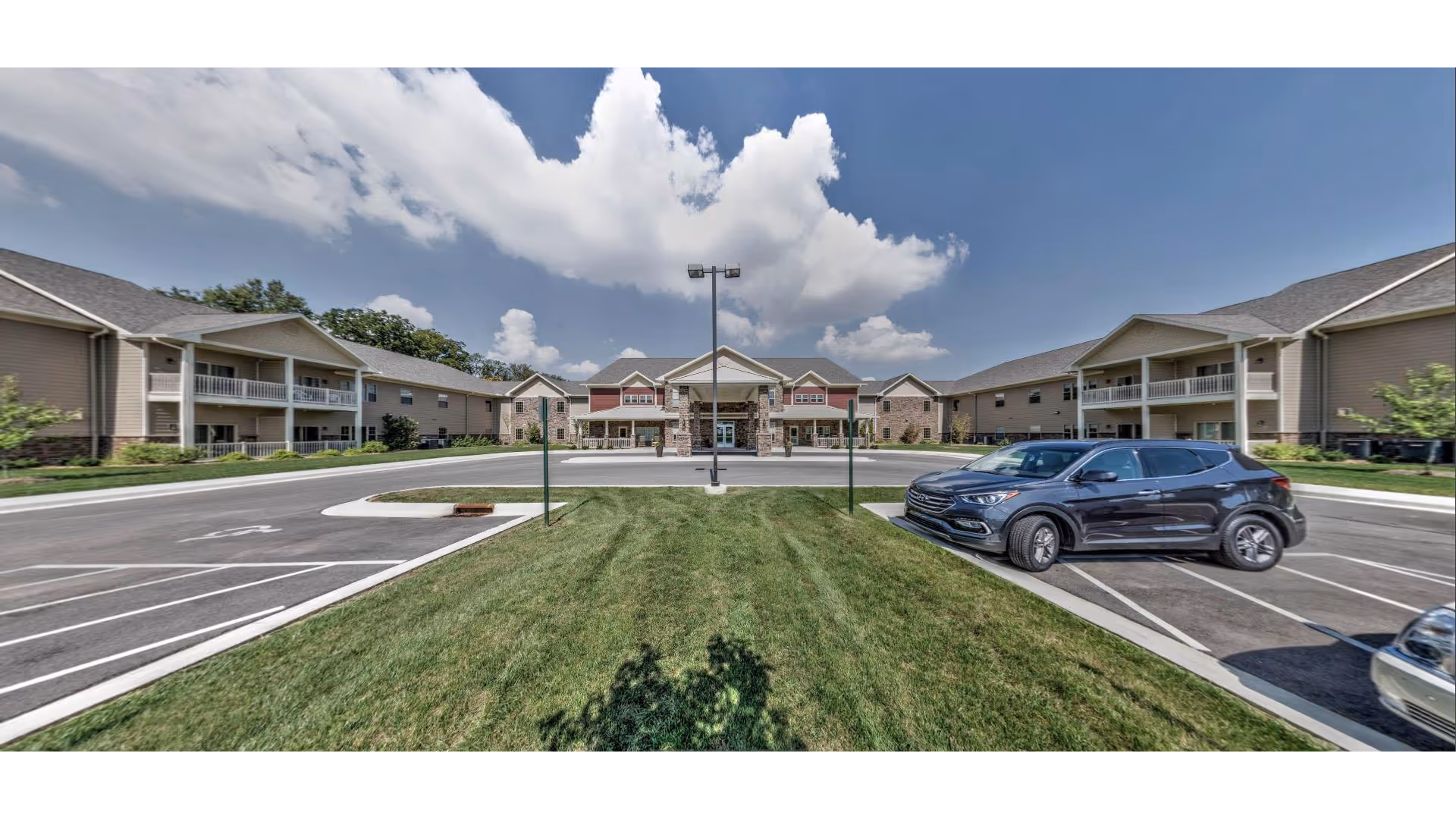 Wide exterior view of Primrose Retirement Community of Newburgh showing a large driveway and parking area with a few cars parked. The building is two stories with balconies and a central entrance under a covered portico. The sky is partly cloudy with bright daylight.