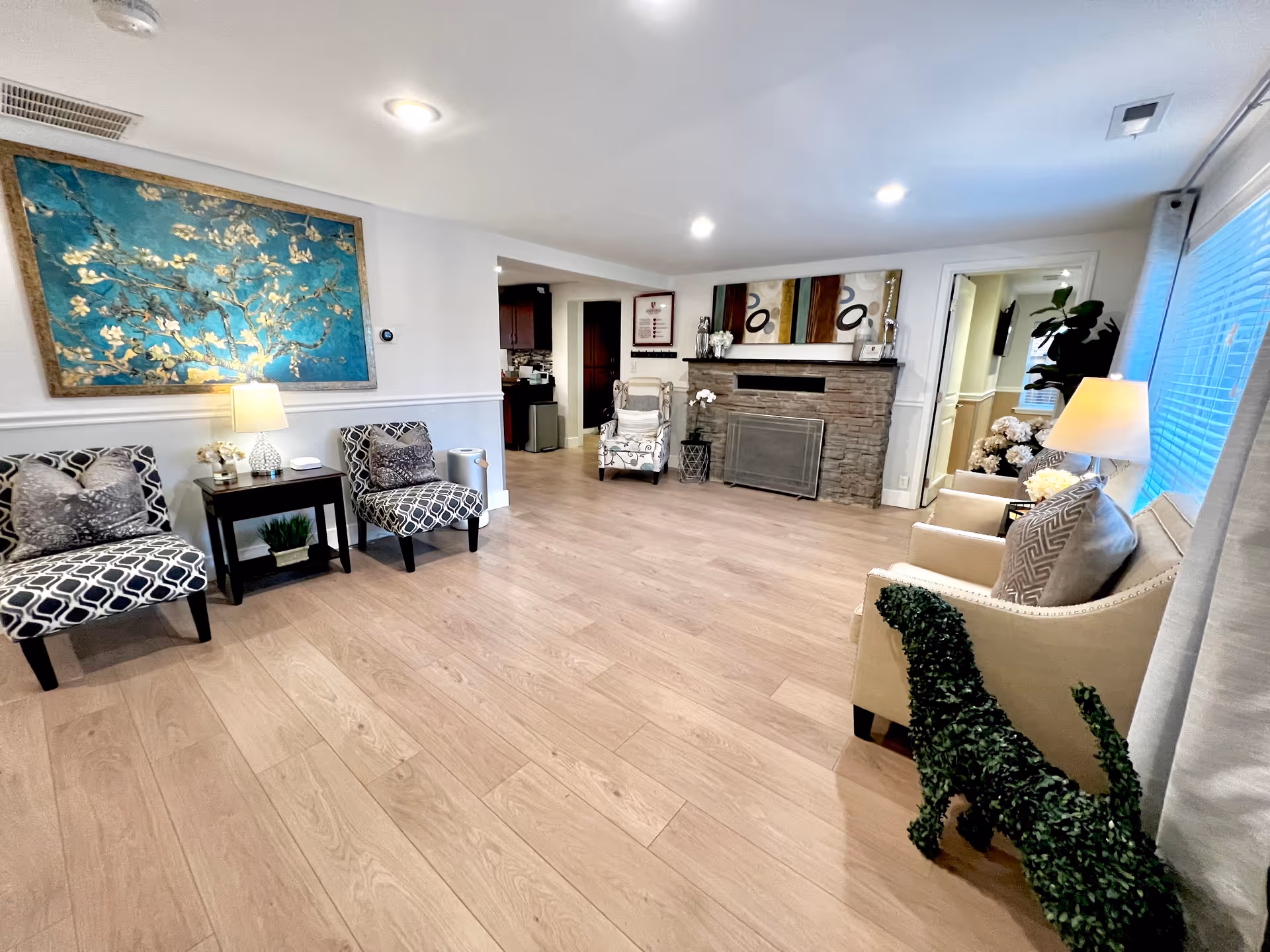 Spacious senior living lounge with patterned chairs, fireplace, artwork, and plants.
