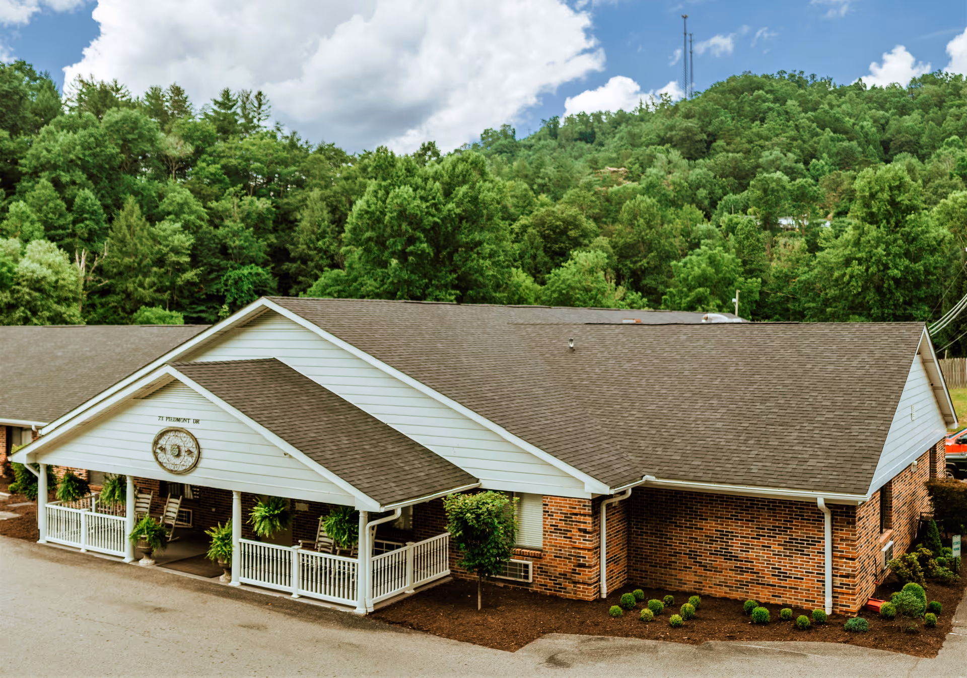 Front exterior of a single-story brick senior living building with a covered porch and wooded hillside behind it.