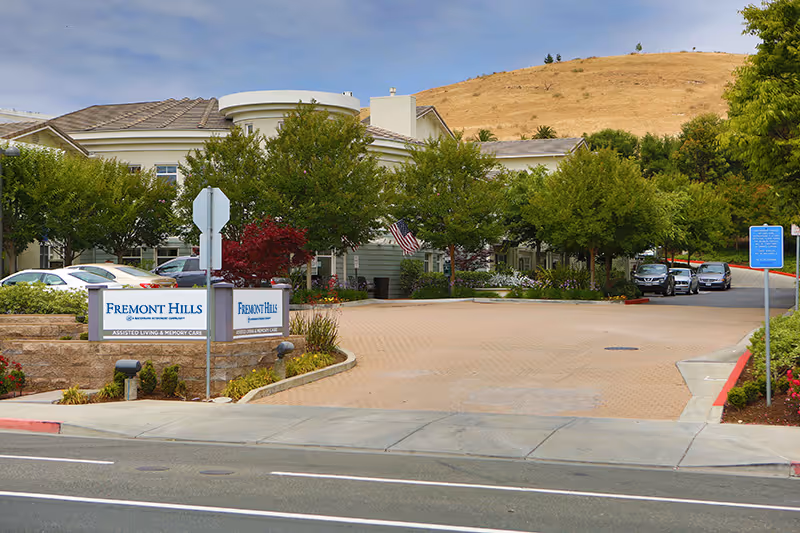 Exterior view of Fremont Hills senior living facility entrance with a driveway, several parked cars, green trees, and a hillside in the background. There are signs displaying the facility name and services offered, including assisted living and memory care.