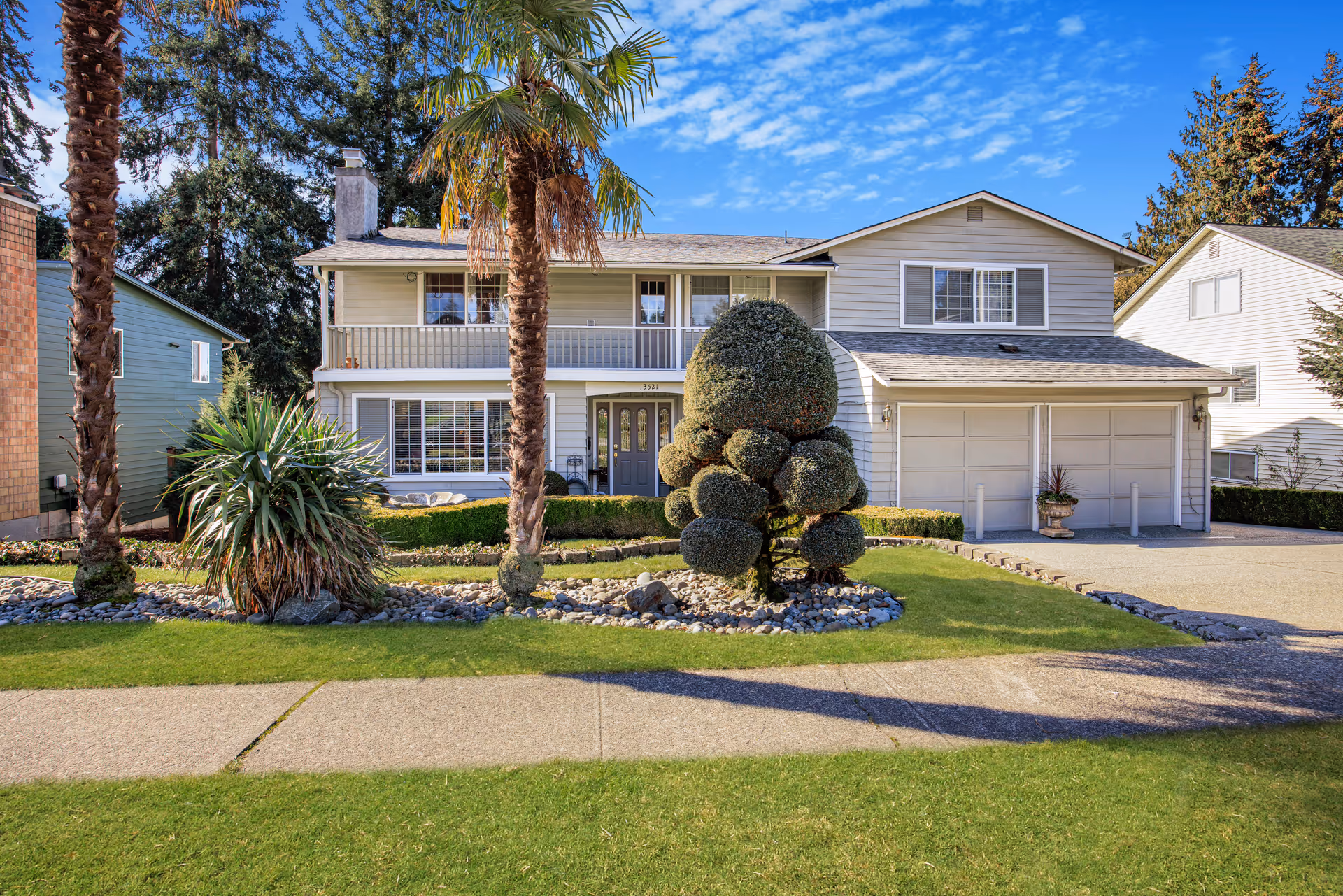 Two-story suburban house with a manicured lawn, palm trees, and decorative shrubbery.