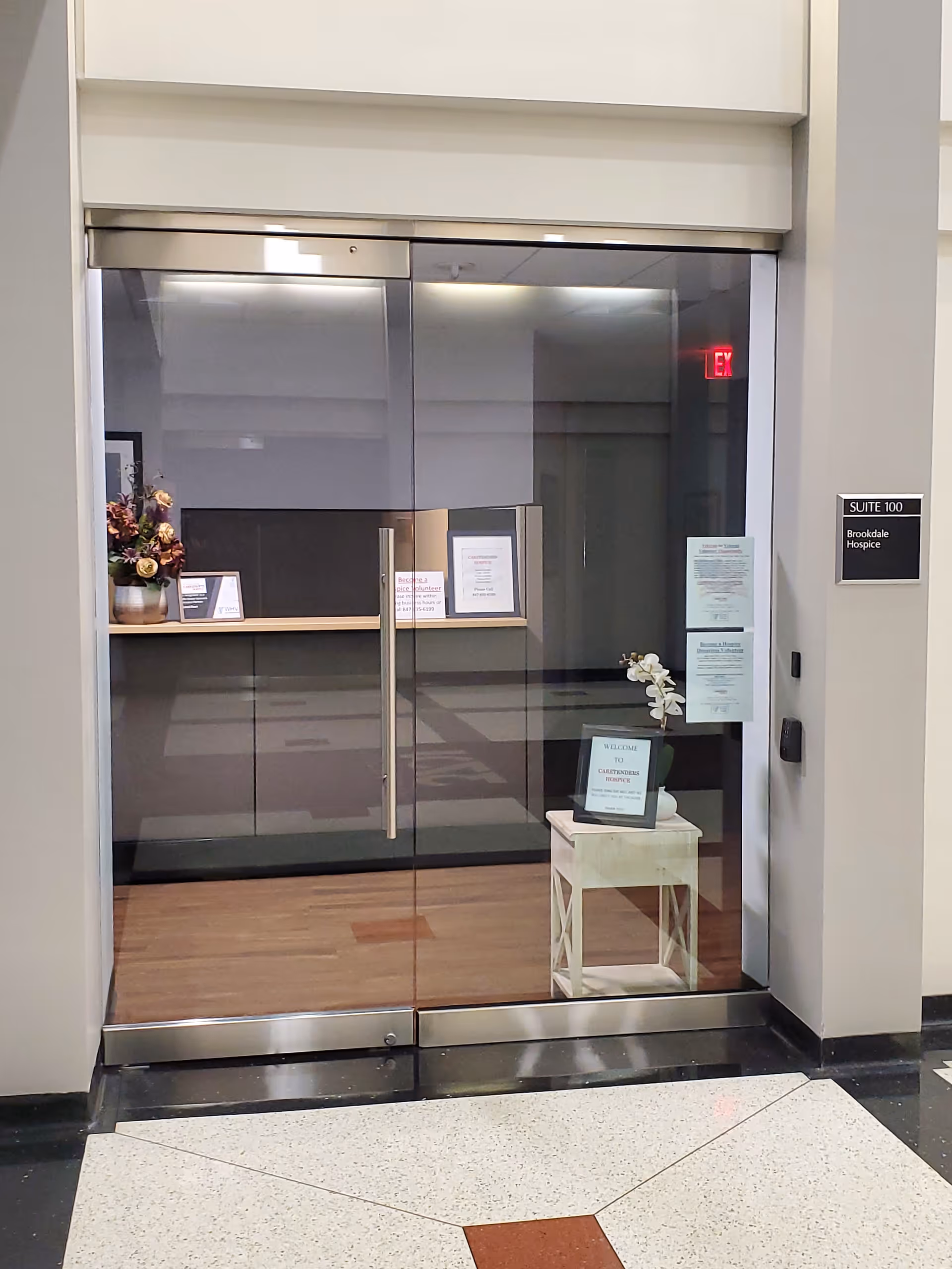 Glass double doors open to a reception area with a small table and signs.