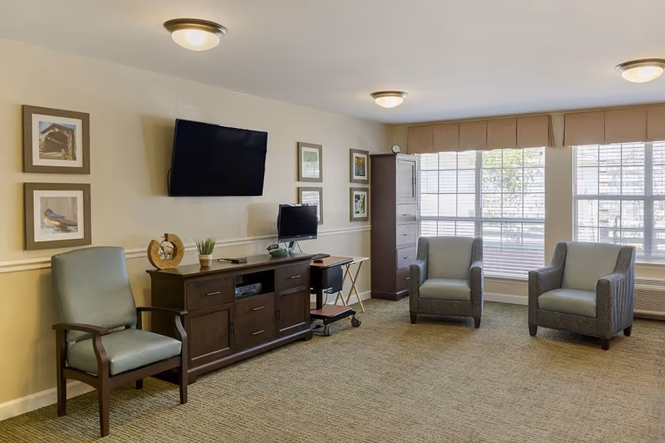 A well-lit living room with three armchairs arranged around a wooden media console. The console holds a small computer monitor and decorative items, and a large flat-screen TV is mounted on the wall above it. The room features large windows with blinds, framed artwork on the walls, and beige carpeting.