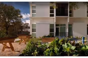 White apartment building with balconies, patio seating, and greenery in the foreground.