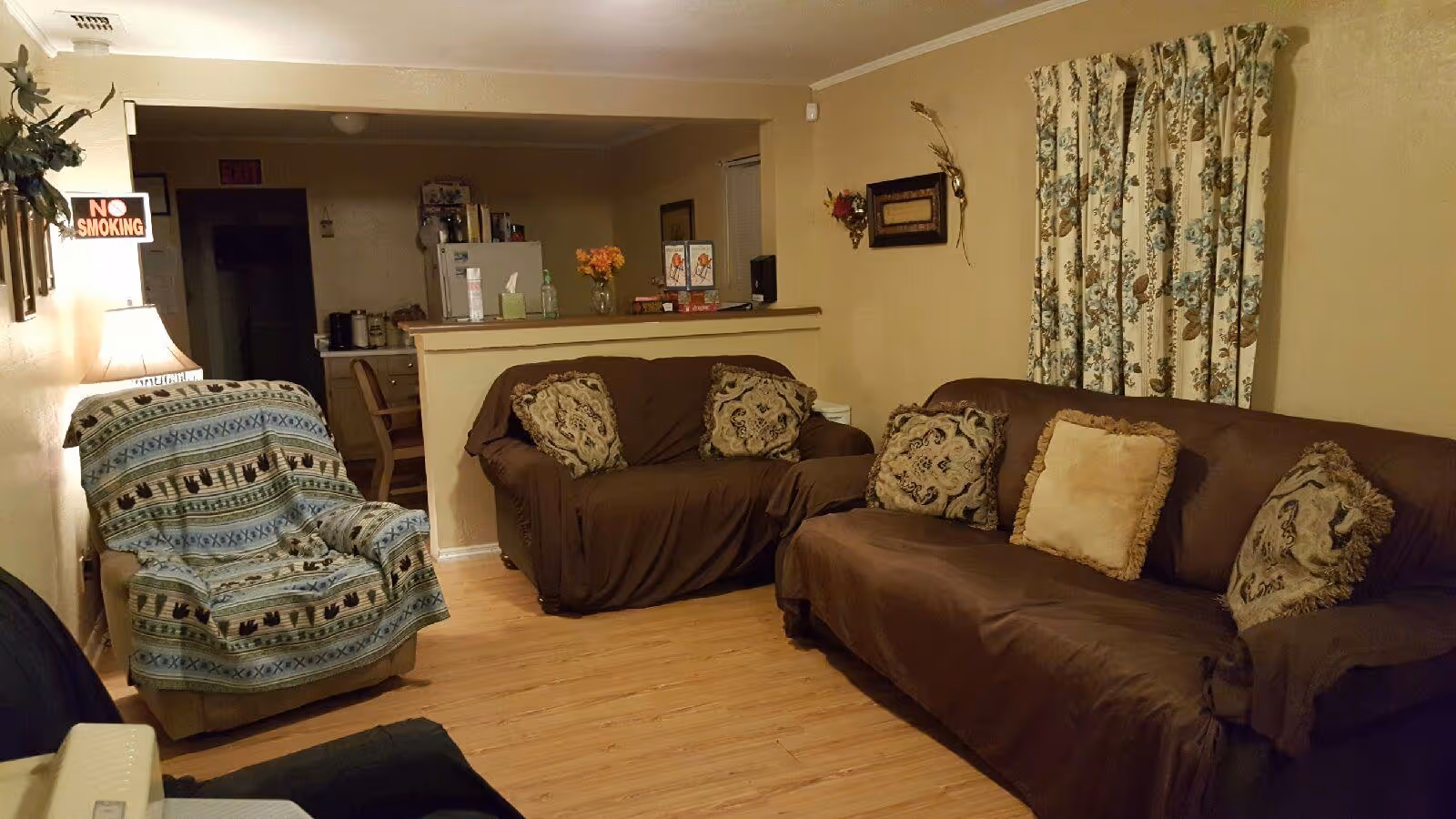 Cozy living room in a senior facility with brown couches, patterned pillows, and floral curtains.